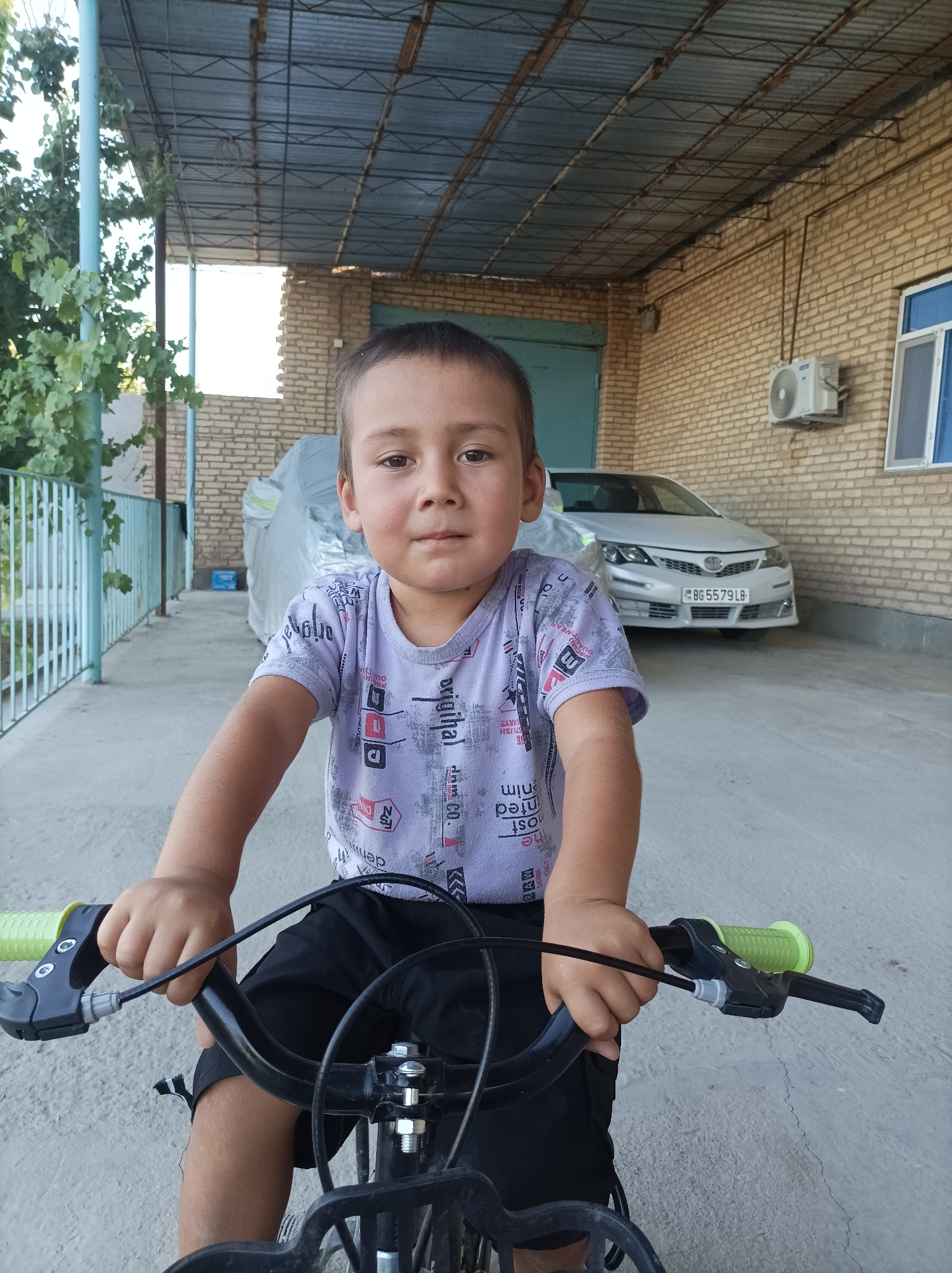 A young boy, likely under ten years old, is positioned on a bicycle, facing forward. He is wearing a light-colored t-shirt with a pattern of text and graphics, and black shorts. His hands are gripping the handlebars of the bicycle, which have bright green grips. The bicycle appears to be black with chrome accents.
The setting is an outdoor, covered area, possibly a carport or a covered patio. The ground is paved concrete. To the left, there is a light blue metal railing and greenery from a tree with leaves. In the background, a light-colored car is parked, with its license plate visible, showing "BG 5579 LB". The car is partially covered with a light gray material. Behind the car, a brick wall is visible, constructed from light brown bricks. Mounted on the brick wall is a white air conditioning unit. To the right of the car and brick wall, a window with blue trim is present. A corrugated metal roof is overhead, supported by metal beams. The overall impression is of a residential outdoor space. The image appears to have been taken during daylight hours, with the sun casting shadows. The location is identified as Seýdi, Turkmenistan.