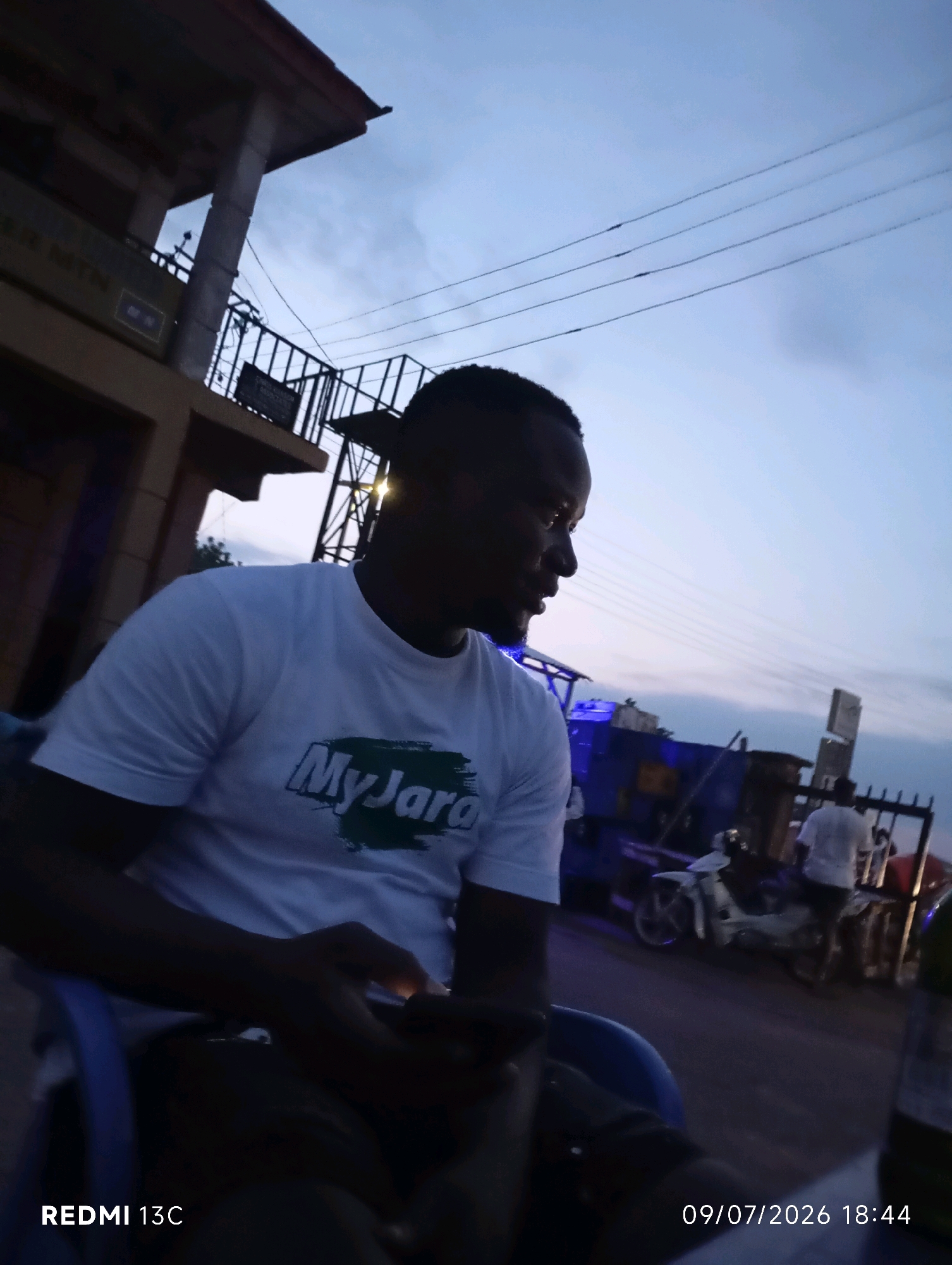 The image depicts an outdoor scene at dusk in Shendam, Nigeria. The foreground features a man sitting on a blue chair, wearing a white t-shirt with a "MyJara" logo. He is looking to his right, holding a smartphone. In the background, there are motorcycles and several people. A building with signage that partially reads "AM MTN" is visible on the left, and a railing with a sign is to the right. The sky is a twilight blue. The timestamp "09/07/2026 18:44" and "REDMI 13C" are overlaid on the image.