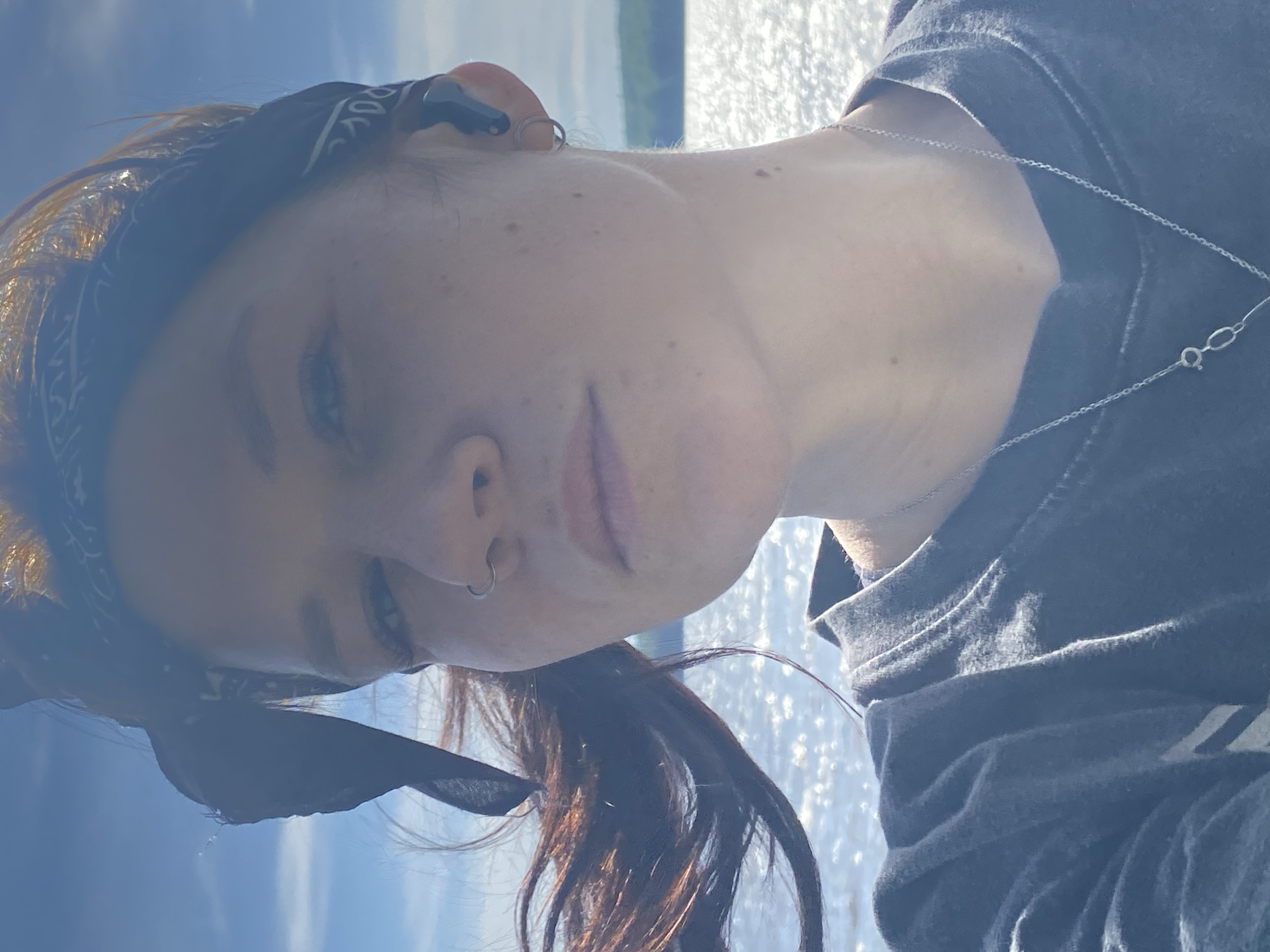 A selfie, taken outside during daytime, shows a person with fair skin, freckles, and reddish-brown hair. They are wearing a dark bandana tied around their head and a dark t-shirt. A silver necklace is visible around their neck. They have a piercing in their left nostril and an earring in their left ear. The background features a body of water with sunlight reflecting on its surface and a treeline in the distance. The location appears to be a natural setting, but it is difficult to pinpoint the specific area in Chaikovsky, Russia without more context.