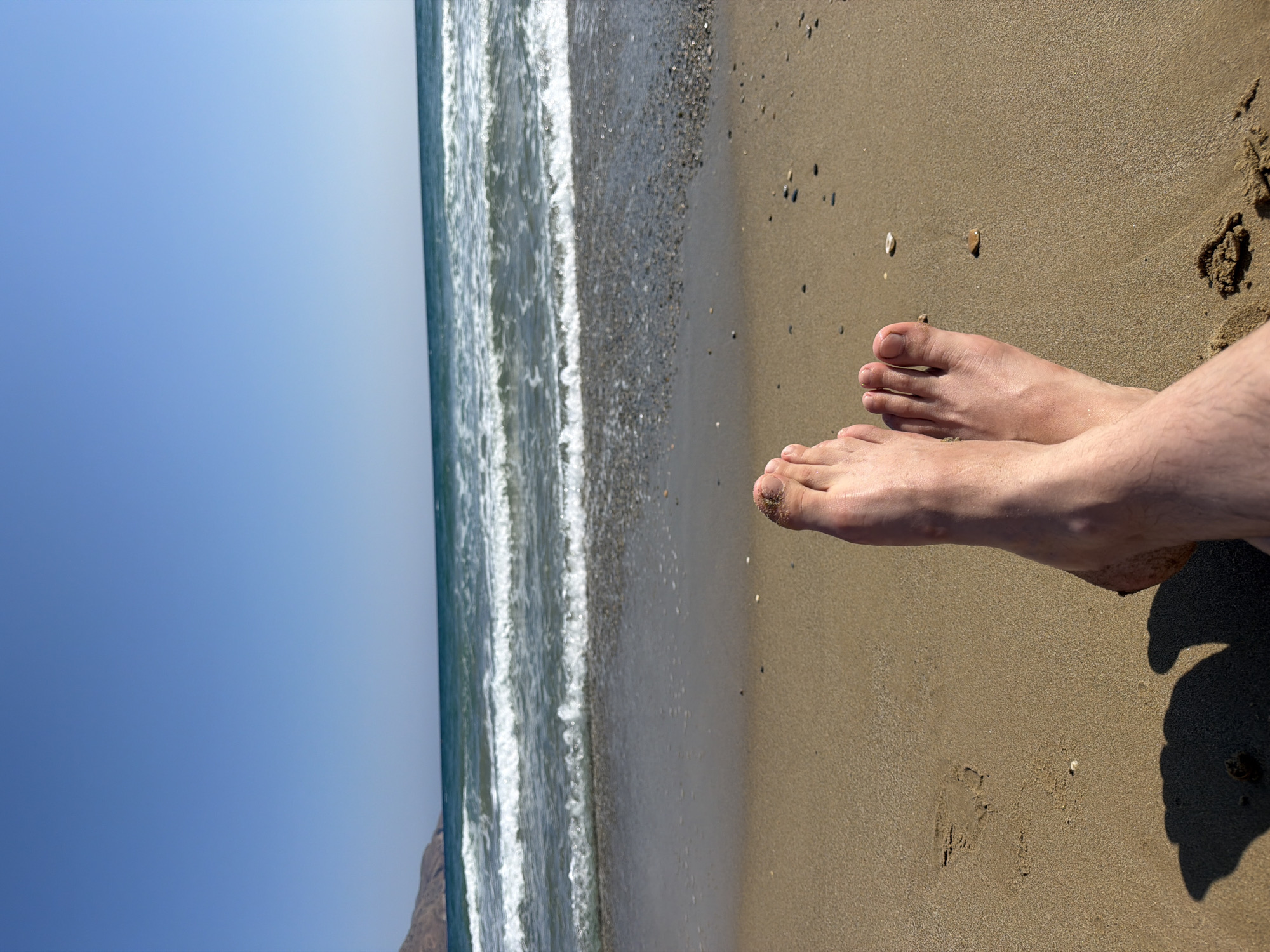 The image shows a pair of bare feet resting on a sandy beach in Georgioupoli, Greece. The toes are pointed towards the viewer, with the left foot slightly overlapping the right. The sand is light brown and appears dry, with a few small pebbles scattered across its surface. In the background, the Aegean Sea is visible with gentle waves rolling towards the shore. The water is a clear turquoise color, and the sky above is a bright, cloudless blue. A faint shadow of the feet and legs is cast on the sand to the left. The scene conveys a sense of relaxation and summer vacation.