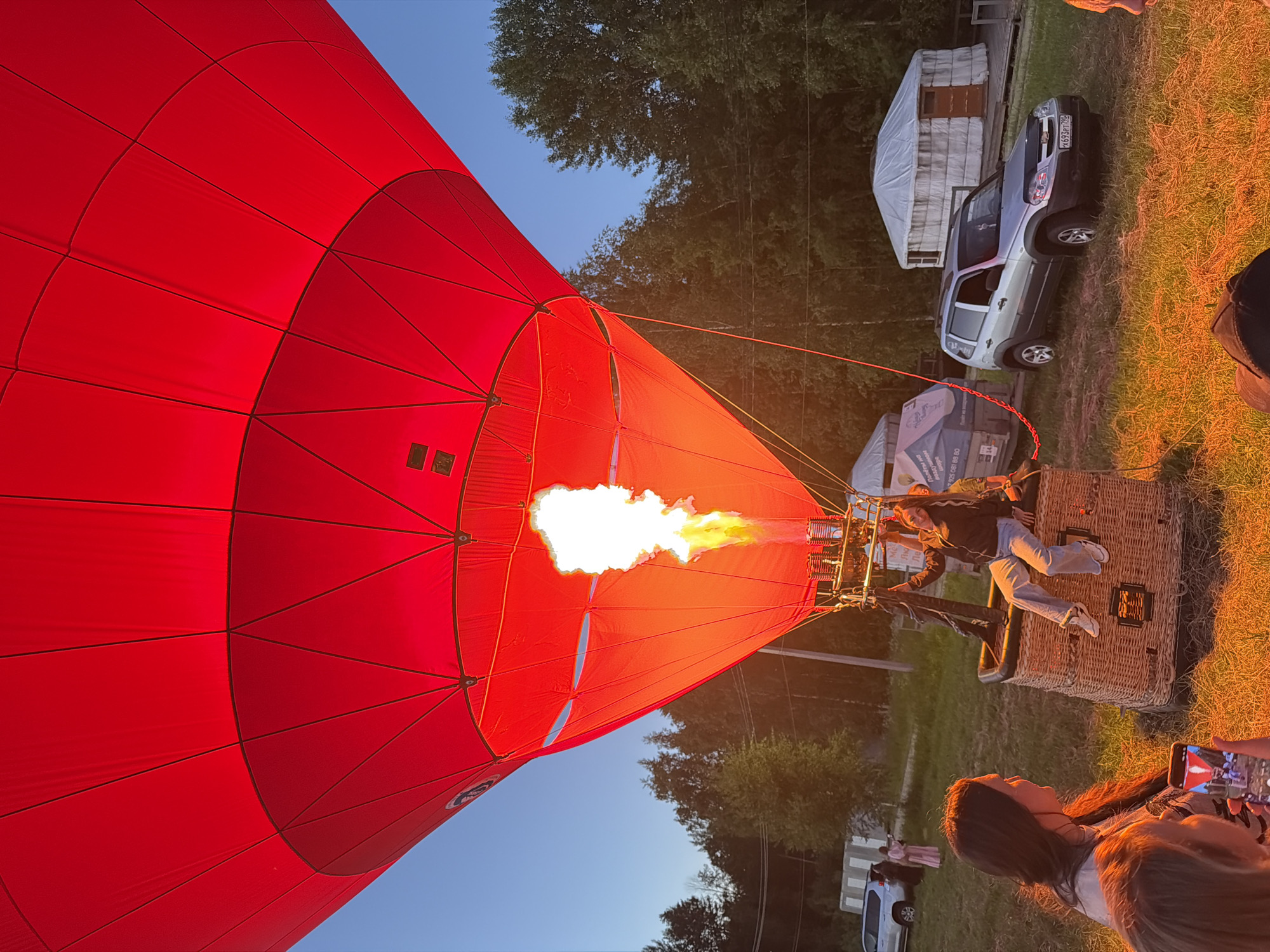 The scene takes place outdoors in a grassy field, likely at dusk, in the city of Unknown, Unknown. A large, bright red hot air balloon is in the process of inflation, dominating the upper and central frame. A powerful, tall yellow-orange flame erupts from the burner assembly directly beneath the balloon's envelope, casting a strong, warm orange glow on the lower part of the balloon, the wicker basket, and the immediate surrounding ground.
In the woven wicker basket of the hot air balloon, a young female with long dark hair, wearing light blue jeans and a dark jacket, is seated on the edge, holding onto ropes and looking slightly to her right. Her feet dangle inside the basket.
In the lower left foreground, a woman with long dark hair, wearing a light-colored top, faces right, observing the balloon. To her left, partially out of frame, another person holds a smartphone horizontally, its screen displaying a live image of the illuminated hot air balloon.
The ground is covered in dry, grassy terrain, vividly lit by the balloon's flame. In the mid-ground, a silver Chevrolet Niva SUV is parked facing slightly right, displaying a Russian license plate "X693PT 199". Behind the SUV, two white, yurt-like tents or structures are visible; one has a wooden door. Further left, a rectangular structure or trailer with faint text is partially obscured. Dense green trees form a backdrop under a clear, light blue sky transitioning to a darker blue at the top, indicative of early evening. Two distant figures are visible near a light-colored vehicle on the far left.