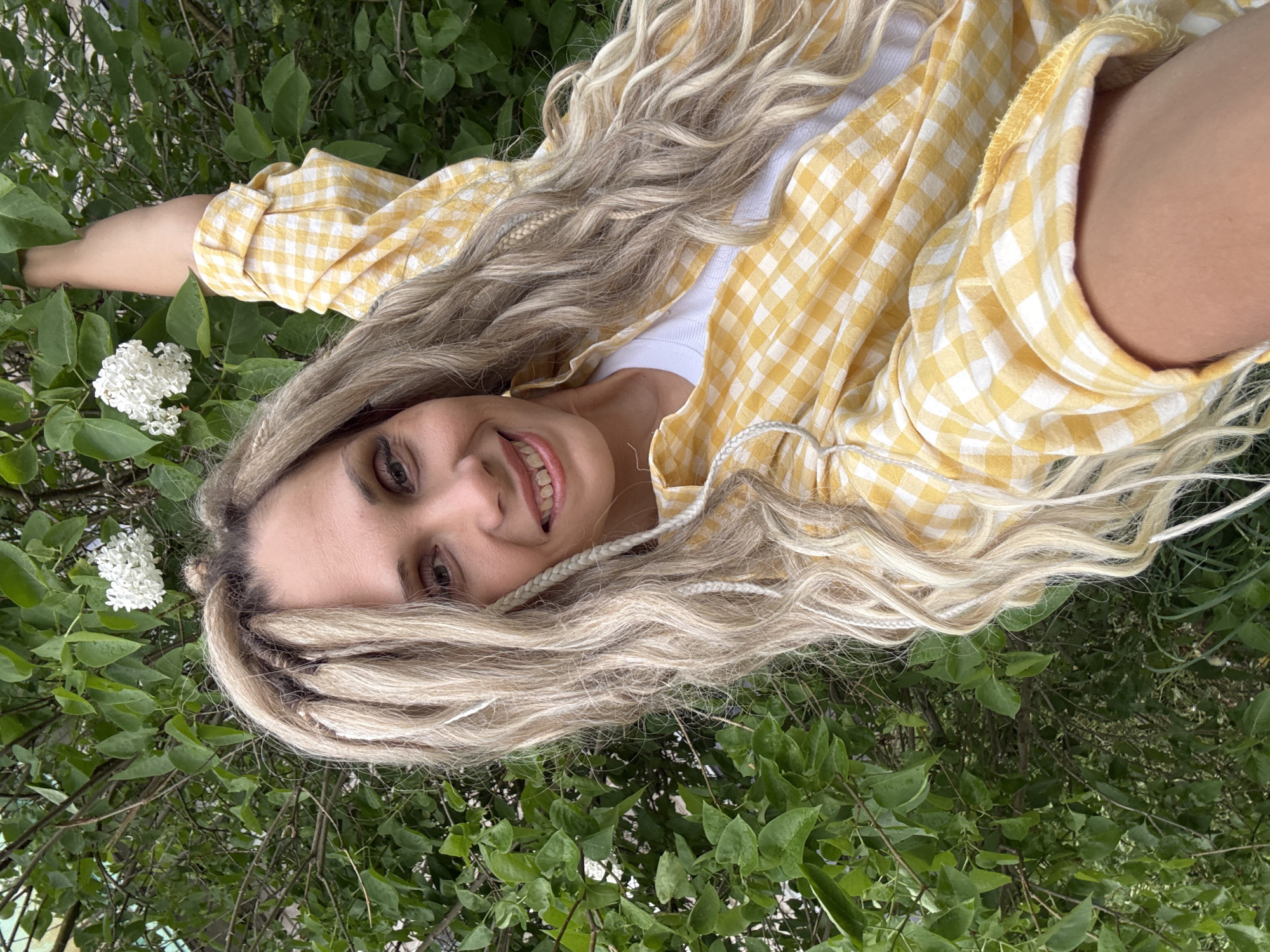 A young woman with blonde hair interwoven with white braids lies on her back amidst lush green foliage. She is wearing a white t-shirt beneath a yellow and white gingham patterned shirt. Her head rests near a cluster of white flowers. The environment appears to be outdoors, possibly a garden or park in Rusanovo, Russia.