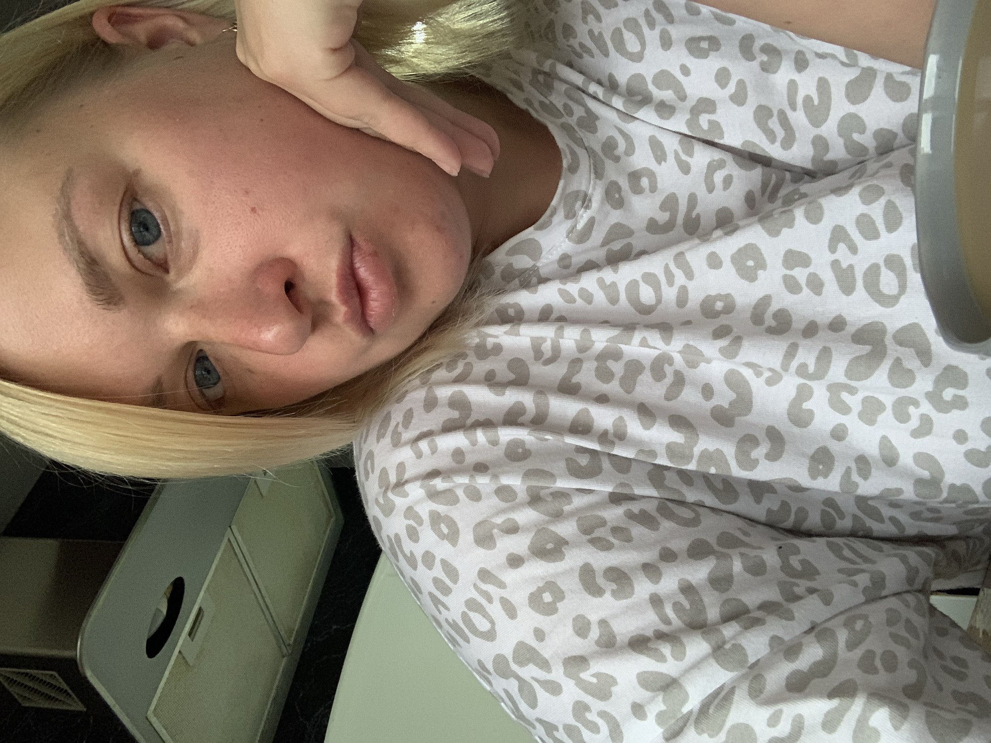 A fair-skinned woman with blonde hair and blue eyes is positioned at an angle in the frame, with her face resting on her hand. She is wearing a white, animal-print top. A white or light-colored mug is partially visible on the right. In the background, a kitchen counter and a kitchen appliance, possibly a toaster oven or microwave, can be seen. The image provides no definitive evidence to suggest a location in the city of Chaikovsky, Russia.