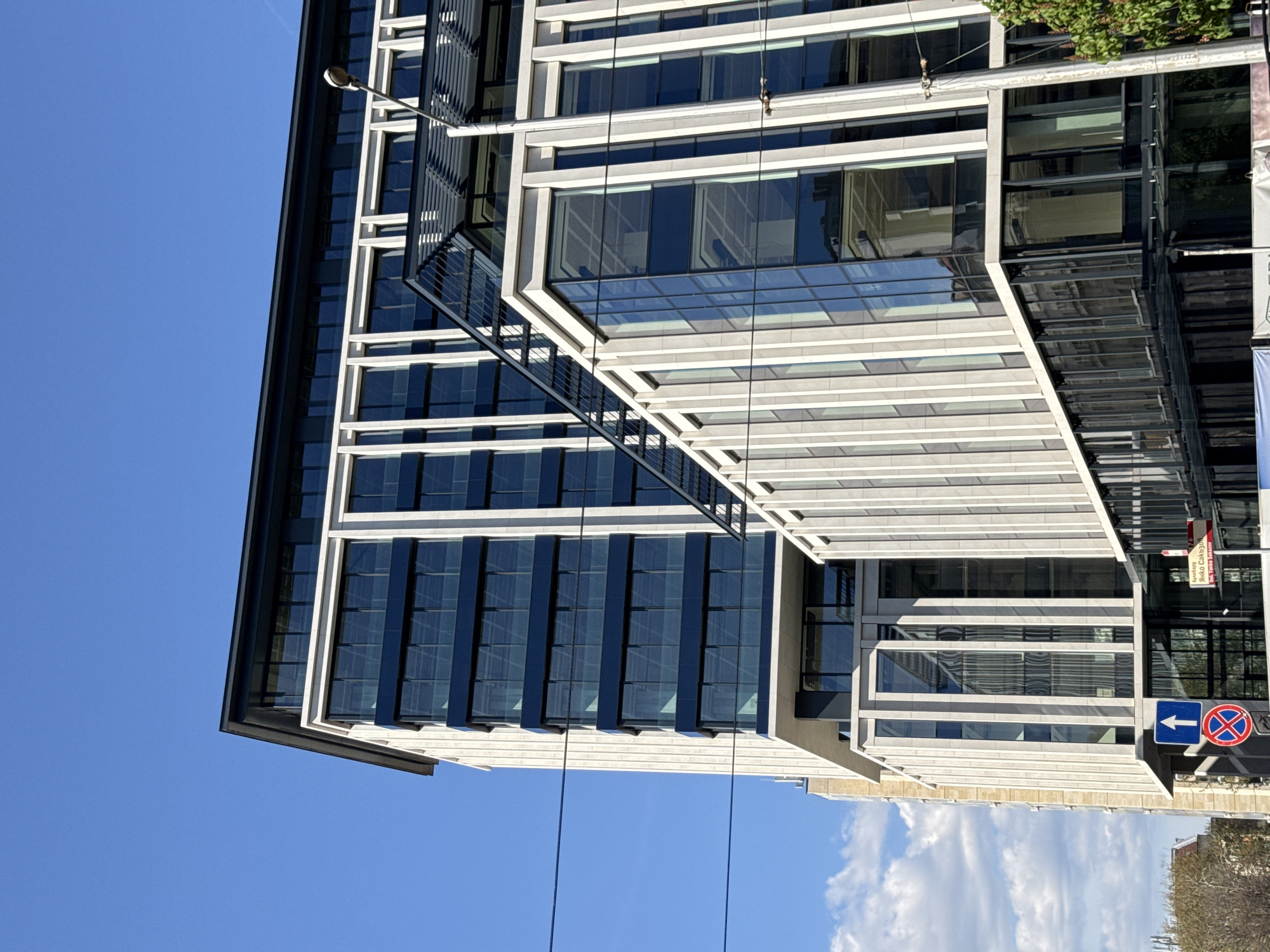 The image displays a modern building in Sofia, Bulgaria, under bright daylight with a clear blue sky and scattered white clouds. The building features a contemporary architectural style, characterized by a facade of large, dark glass panels interspersed with light-colored concrete or stone framing. A distinctive element is an angled section with horizontal white slats, creating an overhang structure that adds depth and shadow play.
The time of day is sunny, indicated by the vibrant blue sky and the sharp reflections visible on the glass surfaces of the building. A lamppost extends from the top of the building, reaching towards the sky. Several thin black utility cables crisscross the foreground against the building and sky.
In the lower right corner, there are several signs. A blue square traffic sign with a white arrow points to the left. Adjacent to it is a red circular sign with a blue background and a red diagonal line, which typically signifies "No Stopping" or "No Parking." Above these traffic signs, a red rectangular sign is visible, displaying text that reads "ЯКО СОФИЯ" (likely meaning "Awesome Sofia" or "Cool Sofia") and "БЪЛГАРИЯ" (Bulgaria). Green foliage is partially visible at the top right edge, suggesting the presence of nearby trees or plants.
The scene is static, focusing on the architectural details and the interplay of light and shadow, with no apparent activity or people.
