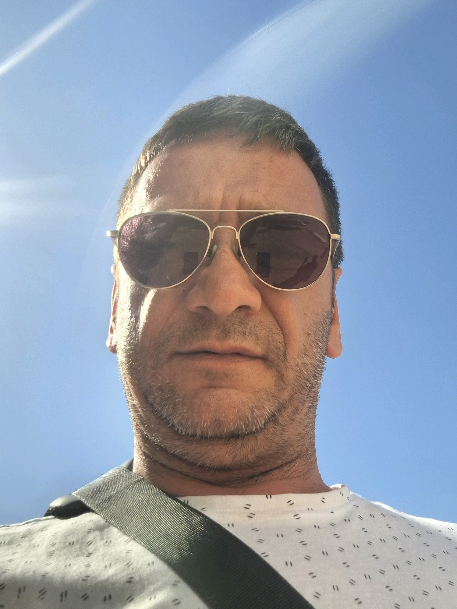 A man wearing sunglasses and a white t-shirt with a pattern of small, dark dashes is shown from the chest up, against a bright blue sky with some lens flare. He has short, dark hair with some graying at the temples and a stubbly beard. A dark strap is visible across his left shoulder. The image appears to be a selfie taken outdoors. There is no visible indication of the city of Kiryat Gat, Israel.