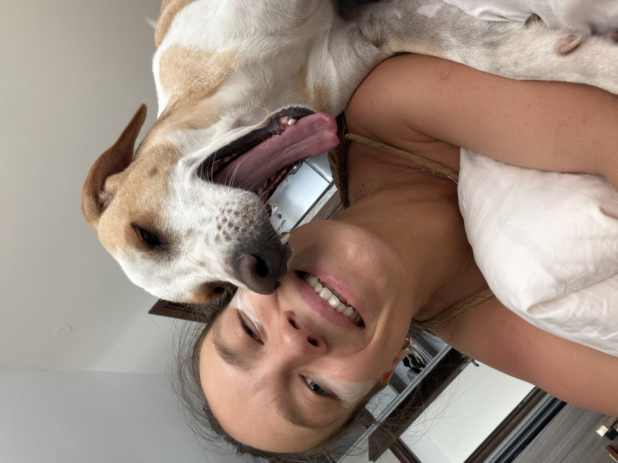 This is a close-up, indoor selfie taken in Budva, Montenegro. A young woman with dark hair, wearing an eye mask and a light-colored tank top, is lying in bed. Her mouth is open in a wide smile, and her eyes are looking towards the camera. A light-colored dog with brown markings is leaning over her, with its mouth open as if yawning or licking her face. The dog has its tongue extended, and its teeth are visible. The background appears to be a bedroom with a white wall and a hint of a closet on the left. The overall impression is one of playful intimacy between the woman and her dog.