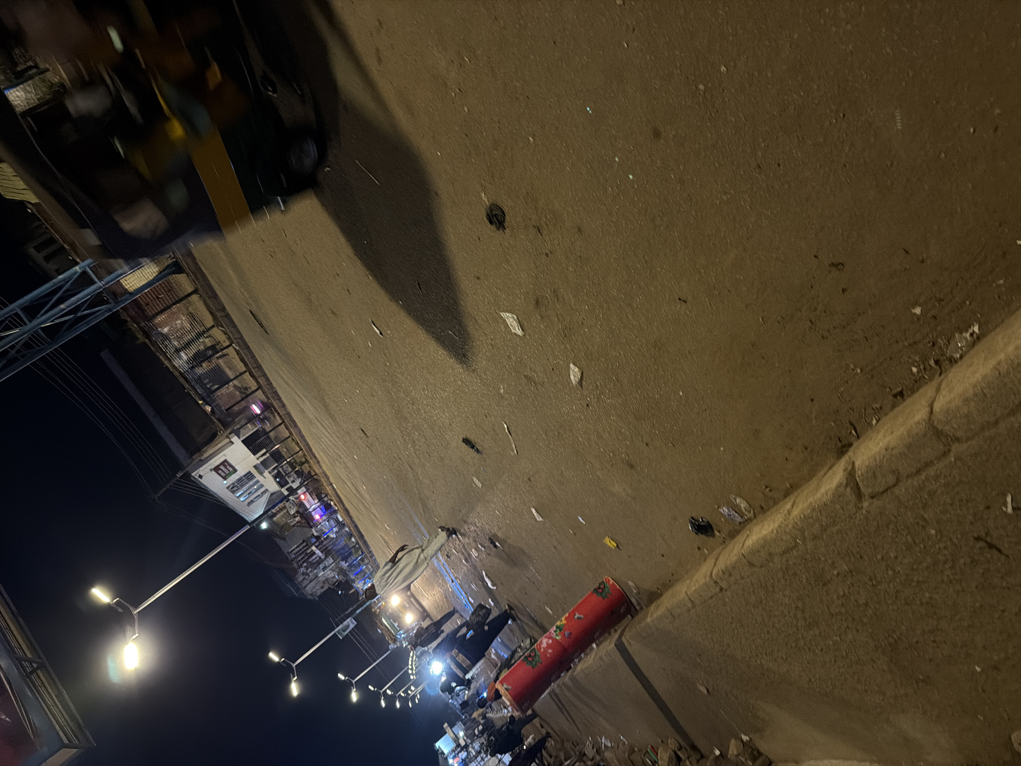 The image shows a nighttime street scene in Bukuru, Nigeria. A red, cylindrical object with a floral pattern is prominently placed on the left side of the frame, near a raised curb. The street itself appears somewhat dirty, with scattered pieces of paper and debris.
Several people are visible on the street. Some are walking, while others appear to be standing or interacting. One person in the foreground is wearing a light-colored robe. In the background, a three-wheeled vehicle, possibly a tuk-tuk, is moving, and its headlights illuminate the road. Streetlights are also visible, casting light on the scene. Buildings line the street, some with illuminated windows. The overall impression is that of a busy, though possibly slightly disorganized, urban environment at night.