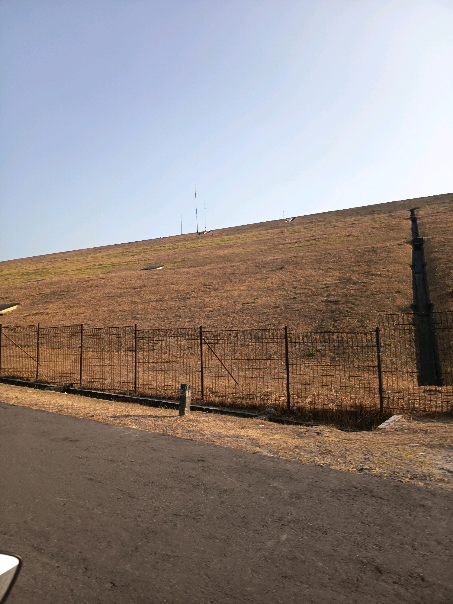 The image depicts a vast, grass-covered embankment with a clear blue sky overhead. A metal fence runs along the base of the embankment, separating it from a paved road in the foreground. There are several dark rectangular structures visible on the embankment, and a dark, vertical pipe-like structure is also present on the right side of the slope. Three thin, vertical poles are spaced out across the top of the embankment, possibly serving as communication towers or markers. The ground on the embankment appears dry and mostly covered in sparse brown grass, with patches of green. The road in the foreground is asphalt and shows some wear. The overall scene suggests an outdoor, possibly rural or undeveloped, location.