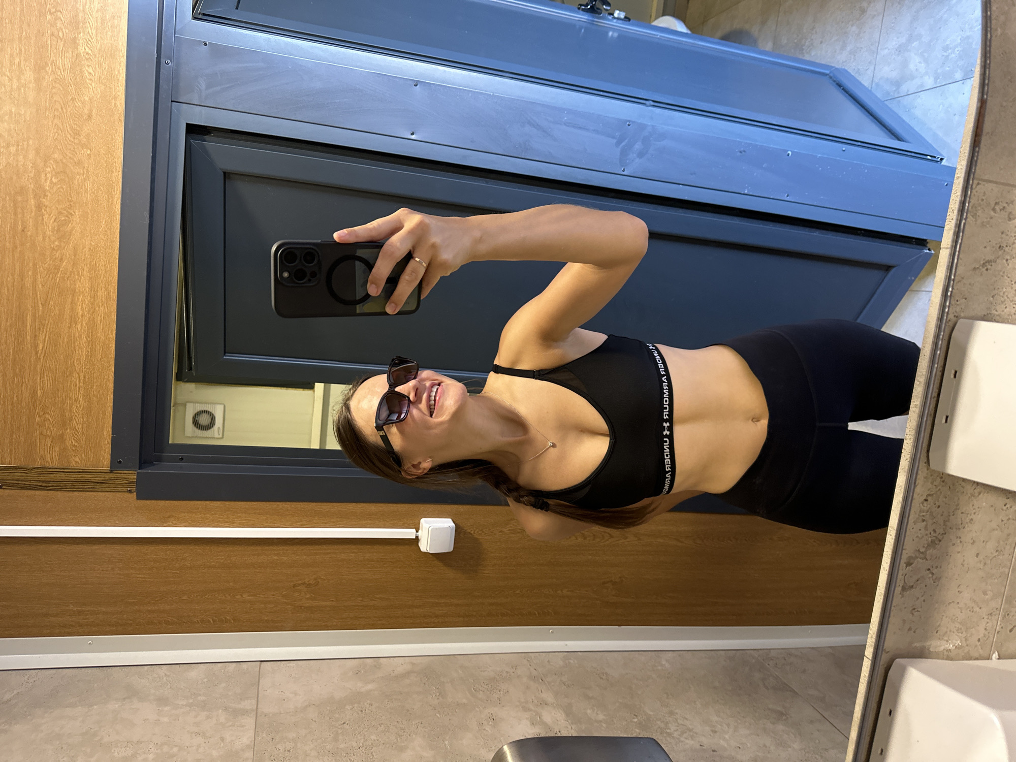 A woman with brown hair in a braid and sunglasses is taking a mirror selfie in what appears to be a locker room or restroom. She is wearing a black sports bra and black leggings. The background shows wooden panels, a dark blue door, and a tiled floor. There is a white electrical outlet on the wooden wall. The woman is holding a black smartphone in her right hand and is smiling. The location is specified as Boreti, Montenegro.