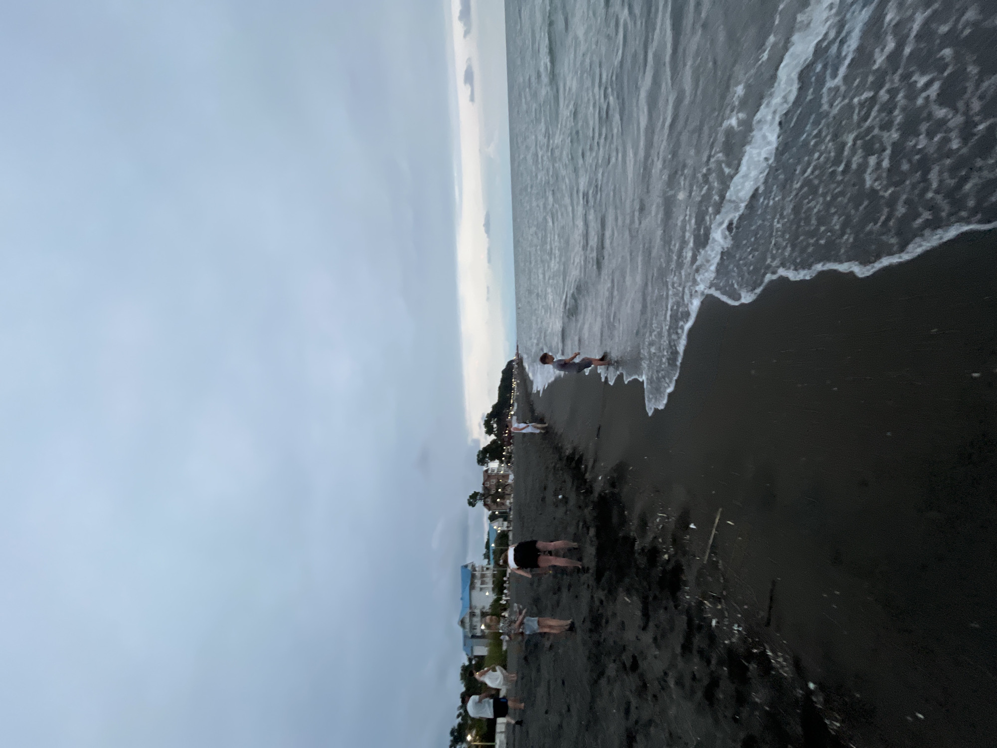 The image depicts a wide, dark sand beach in Ureki, Georgia, under a cloudy, twilight sky. The sea, with gentle waves, meets the shore on the right side of the frame. Several people are scattered across the beach, some walking and others standing near the water's edge. In the background, along the coast, a row of buildings, including houses and hotels, are visible, some with illuminated windows. The overall atmosphere is subdued, consistent with the fading light of evening.