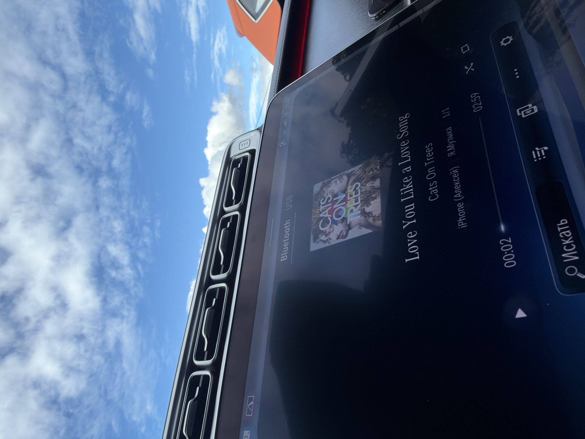 The image shows the interior of a car, specifically the dashboard area. The infotainment screen is displaying the song "Love You Like a Love Song" by Cats On Trees, playing from an iPhone. The time is 16:47, and the song is 2 seconds in with a total of 2 minutes and 59 seconds. To the left of the screen are the air vents of the car. Outside, there is a blue sky with white clouds. Part of a red or orange vehicle is visible in the background, and the photograph was taken inside a Mercedes Benz.