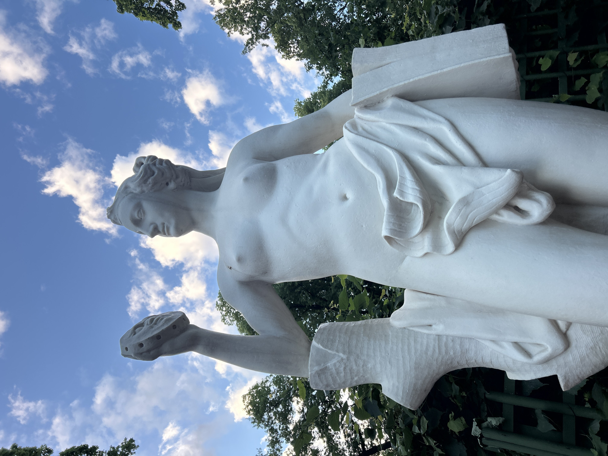 A white marble statue of a nude female figure stands in front of lush green foliage and a blue sky with scattered white clouds. The statue is depicted from the waist up, with her right arm raised, holding a circular object with multiple holes. Her left arm is bent, holding a rectangular object draped with fabric. The statue's head is turned to the left, and she has flowing hair. The base of the statue is partially obscured by a textured, tree-like element on the left. A green metal fence is visible in the background behind the statue. The overall impression is that of classical sculpture set in a natural outdoor environment.