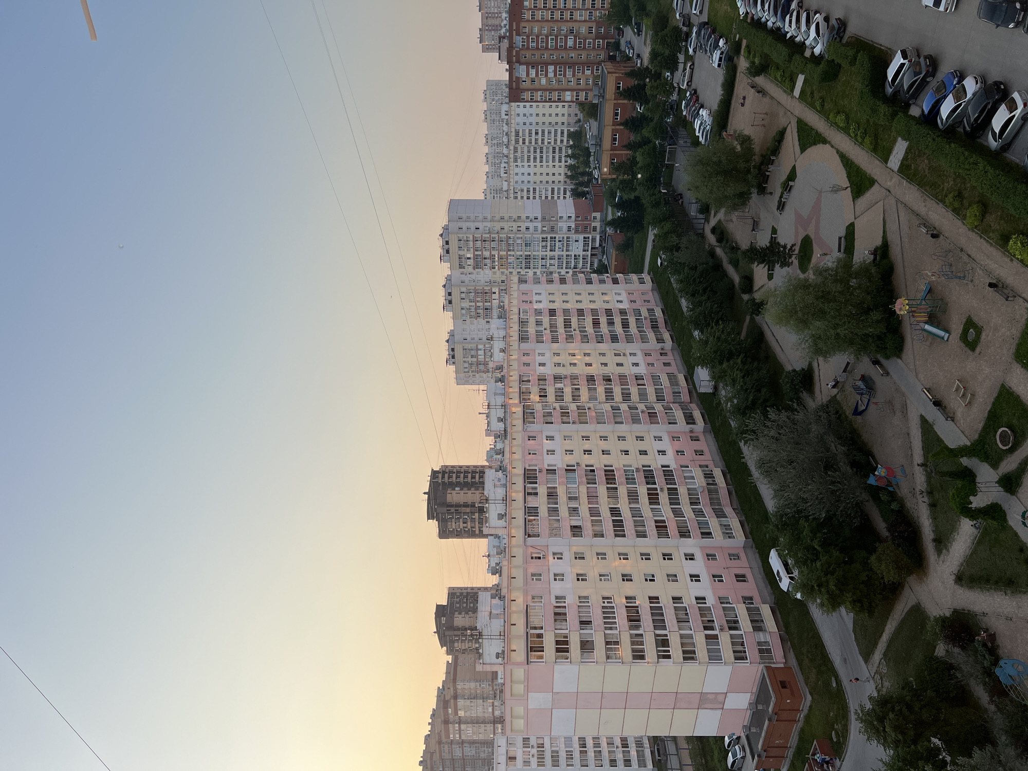 The image depicts a view of a residential area in Novosibirsk, Russia, at dusk. In the foreground, a multi-story apartment building with a pink and yellow facade is prominent. Beyond this building, other apartment complexes of varying heights and architectural styles are visible, with some featuring brick exteriors and others lighter colored facades. The sky is clear with a soft gradient of light blue and pale yellow, indicating sunset or sunrise.
A street runs in front of the apartment buildings, lined with parked cars. A playground area with slides, swings, and sand is situated on the left side of the image, with a few people visible in the vicinity of the playground and walking on a pathway. Trees and green spaces are interspersed throughout the scene, contributing to a residential park-like atmosphere. Power lines are visible stretching across the sky. The overall scene is typical of a modern urban residential district.