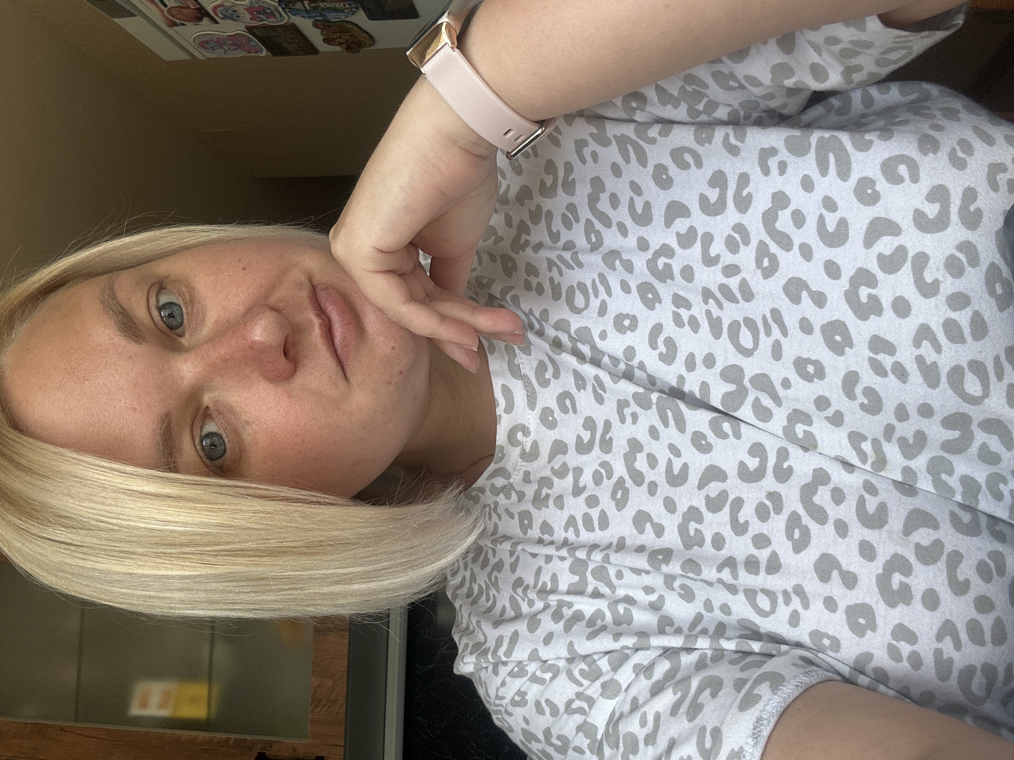 A blonde woman with blue eyes and fair skin is pictured in a close-up selfie. She has her right hand resting on her chin, and she is wearing a light-colored t-shirt with a grey leopard print pattern. A light pink smartwatch is visible on her left wrist. The background of the image is slightly blurred, showing kitchen cabinets and a refrigerator with stickers. The lighting suggests an indoor setting, possibly a kitchen. The image does not contain any specific visual cues or landmarks that would place it in the city of Chaikovsky, Russia.