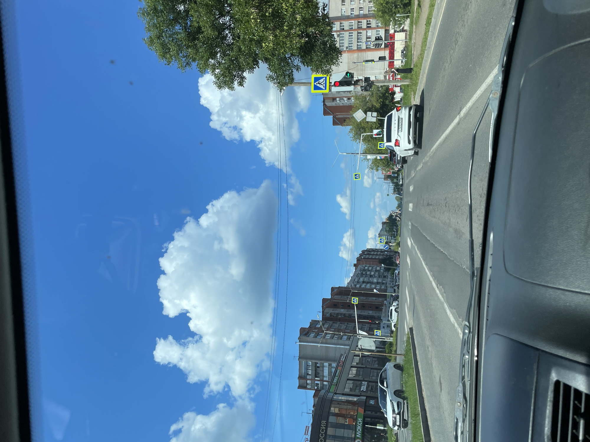 The image captures a view from inside a vehicle driving on a sunny day in Kirovo-Chepetsk, Russia. The scene depicts a city street with multi-story apartment buildings lining both sides. The road has multiple lanes and is marked with white lines. Pedestrian crossing signs are visible at intersections. Several cars are driving on the road, and some are parked. A large green tree is on the right side of the image. The sky is bright blue with scattered white clouds.