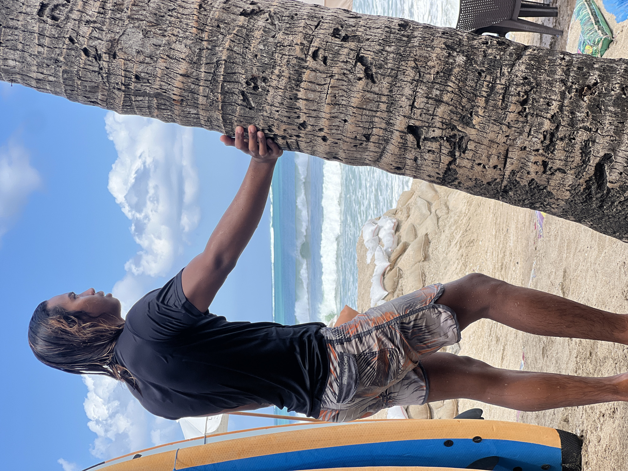 A male surfer with long, wet hair stands on a sandy beach, his back to the camera, looking out at the ocean. He is wearing a black rash guard and patterned swim shorts. A surfboard, predominantly orange and blue, leans against a palm tree to his left. The palm tree's trunk is textured and rough. In the background, the ocean has moderate waves, and sandbags are piled on the shore, likely for erosion control. The sky is blue with scattered white clouds. The scene appears to be the Kuta beach in Bali, Indonesia, a popular destination for surfers.