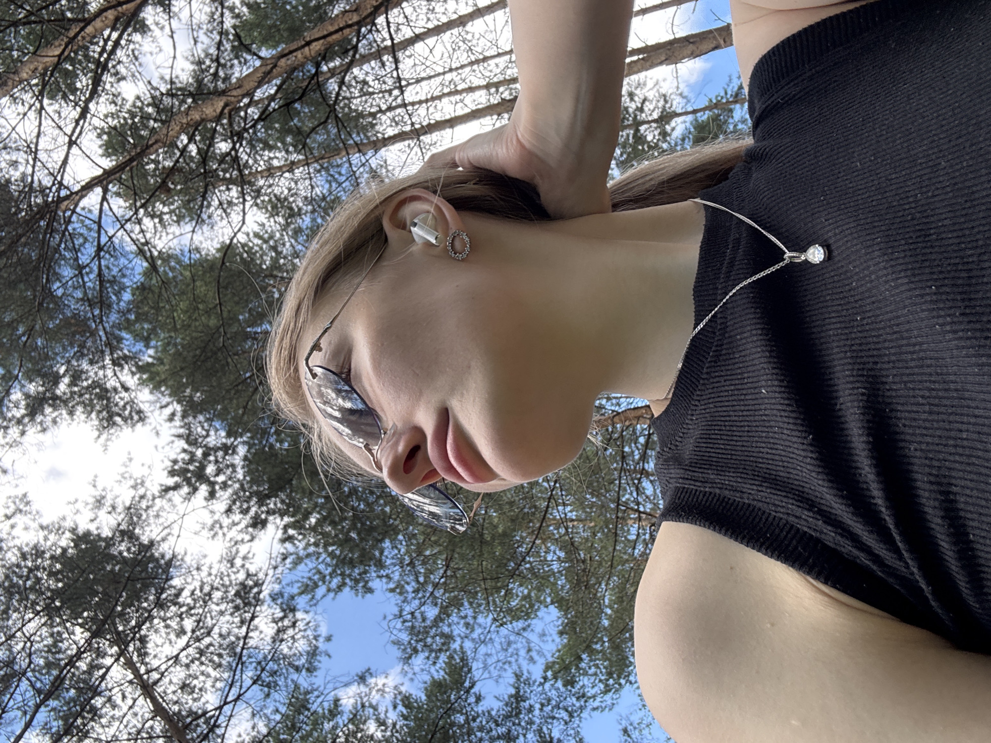 A low-angle, waist-up shot shows a young woman in a black ribbed tank top and sunglasses, looking up and to the left. She has blonde hair pulled back in a ponytail, and is wearing a silver necklace with a teardrop pendant, and a circular earring with small stones. A white earbud is visible in her right ear. Her left hand is resting on the back of her neck. The background consists of a clear blue sky and the tops of tall pine trees, suggesting an outdoor setting in a forest. There is no visible indication of the city of Perm, Russia, or any urban elements.