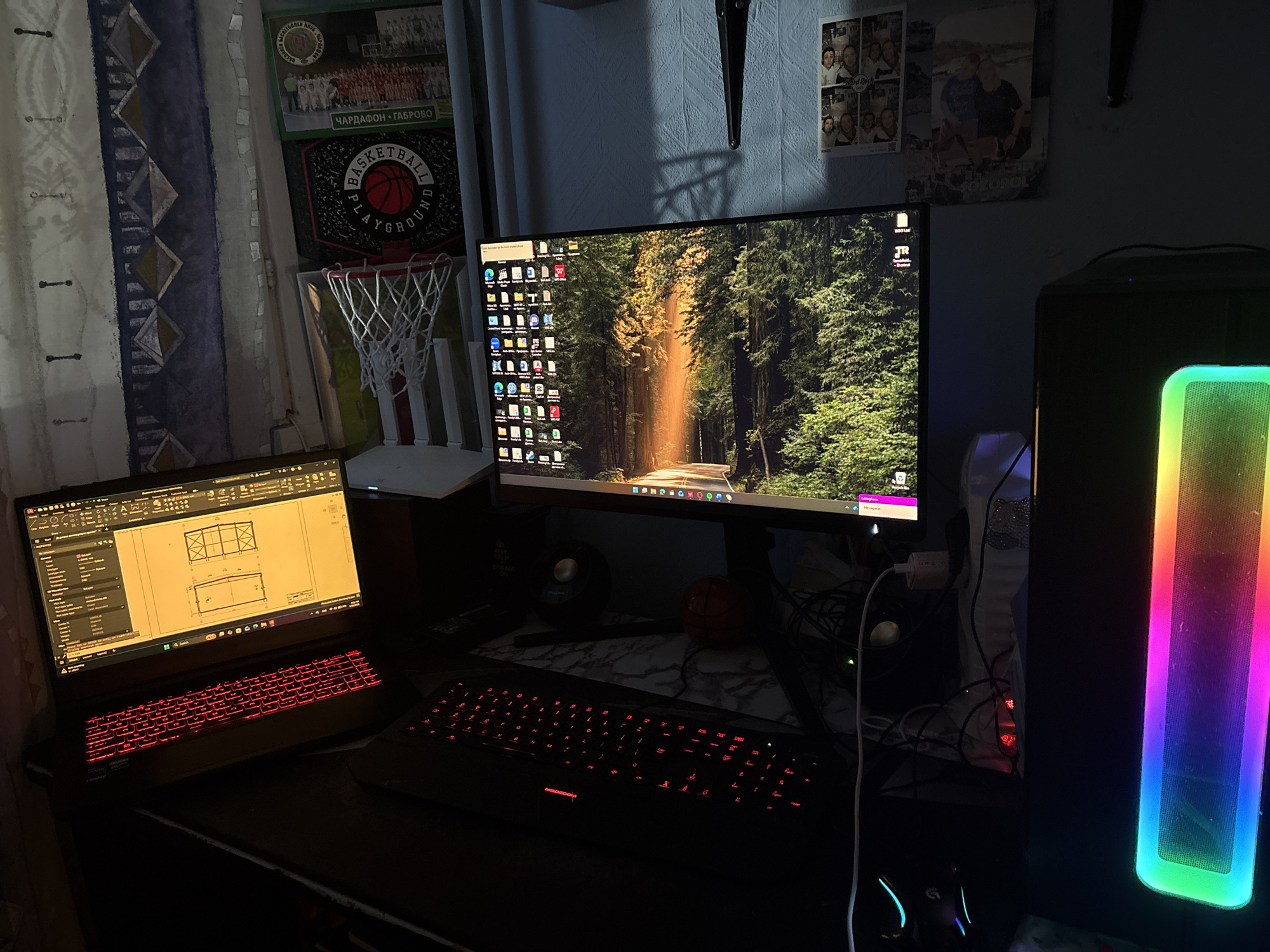 The image captures a dimly lit computer setup in what appears to be a personal space. A laptop on the left displays a technical drawing, its keyboard illuminated in red. A larger monitor in the center shows a nature scene as a desktop background, with numerous icons visible on the left side. A black keyboard sits in front of the monitor. To the right, a desktop computer tower features a vertical, rainbow-colored LED strip. Various cables and peripherals are visible, along with a mouse on the desk. In the background, there's a curtain with a geometric pattern, posters or pictures, including one related to basketball (with the words "BASKETBALL PLAYGROUND"). Another poster seems to say "ЧАРДАФОН - ГАБРОВО" which indicates that this is most likely in Bulgaria. Based on this information, there's no definitive evidence to pinpoint the exact location in Sofia or to say that this is even in Sofia. This looks like a personal space, like a bedroom, and does not provide sufficient information to infer that this is a business in the city of Sofia.