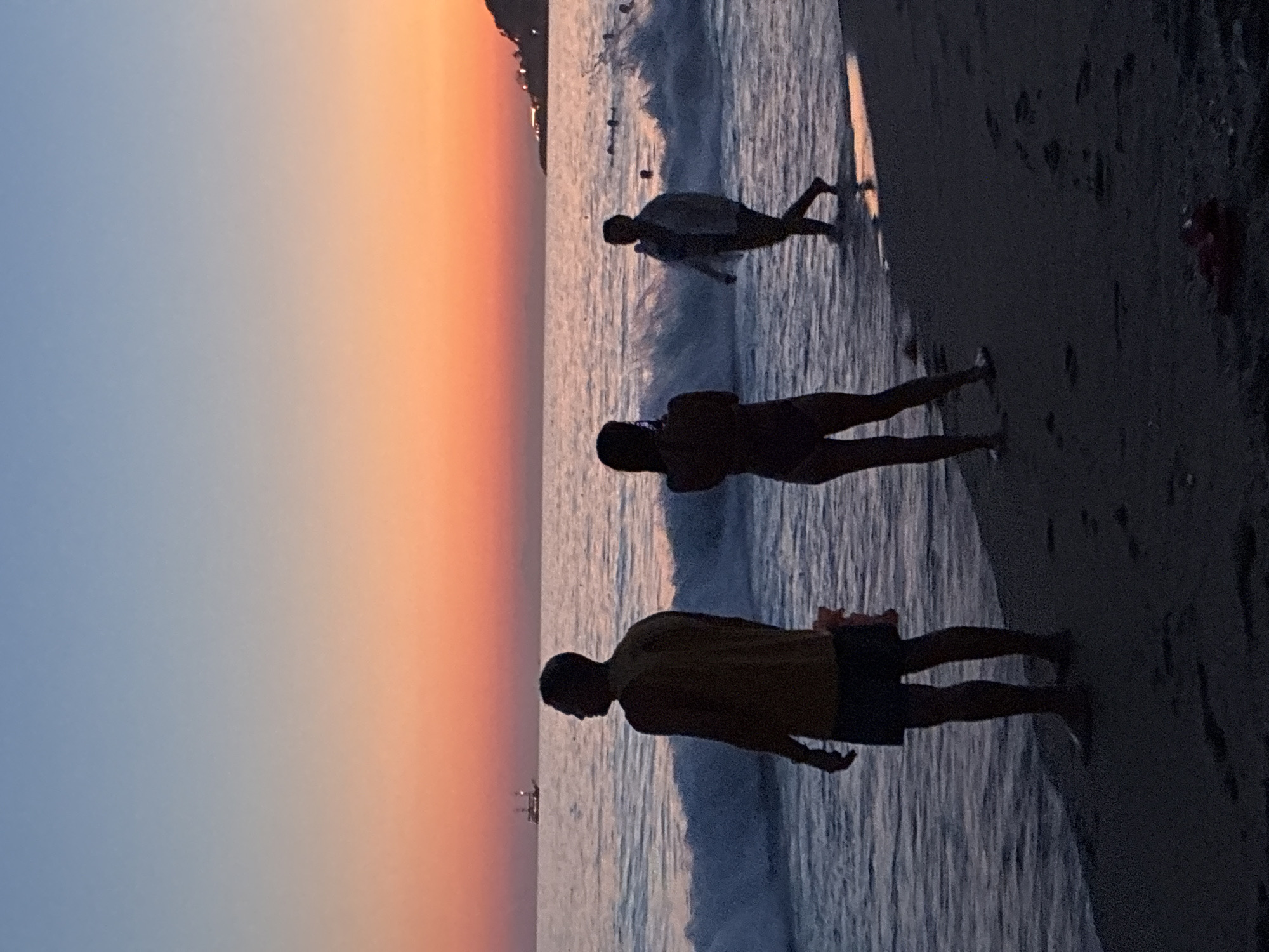 A silhouette of three people on a beach at sunset in Alanya, Turkey. The sky is a gradient of blue and orange. The ocean has gentle waves, and a boat is visible on the horizon. The person in the foreground is a man wearing a yellow shirt and dark shorts, facing away from the camera. To his right, a woman in a swimsuit is taking a picture with her phone. Further in the background, a person in a light-colored jacket walks into the water. Some people are swimming in the distance. The sand on the beach is dark, with footprints visible. A red flip-flop is on the sand in the lower right corner.