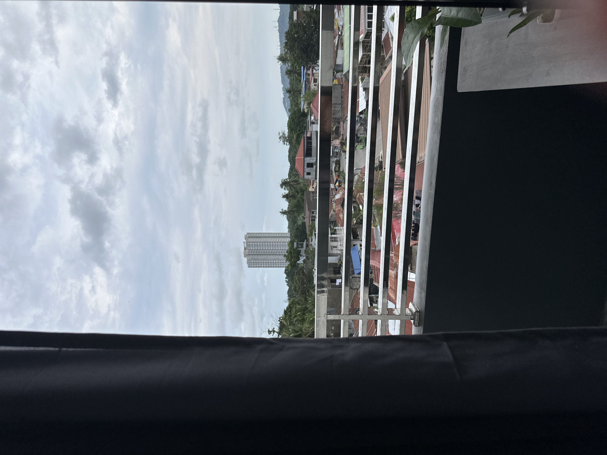 The image depicts a view from a window or balcony in Davao City, Philippines. In the distance, a tall condominium building is visible, surrounded by trees and smaller houses. In the midground, there are houses with red corrugated metal roofing. A silver-colored metal balcony railing is in the foreground. The sky is overcast with clouds. The view is partially obstructed by a dark gray curtain or fabric along the bottom and right side.