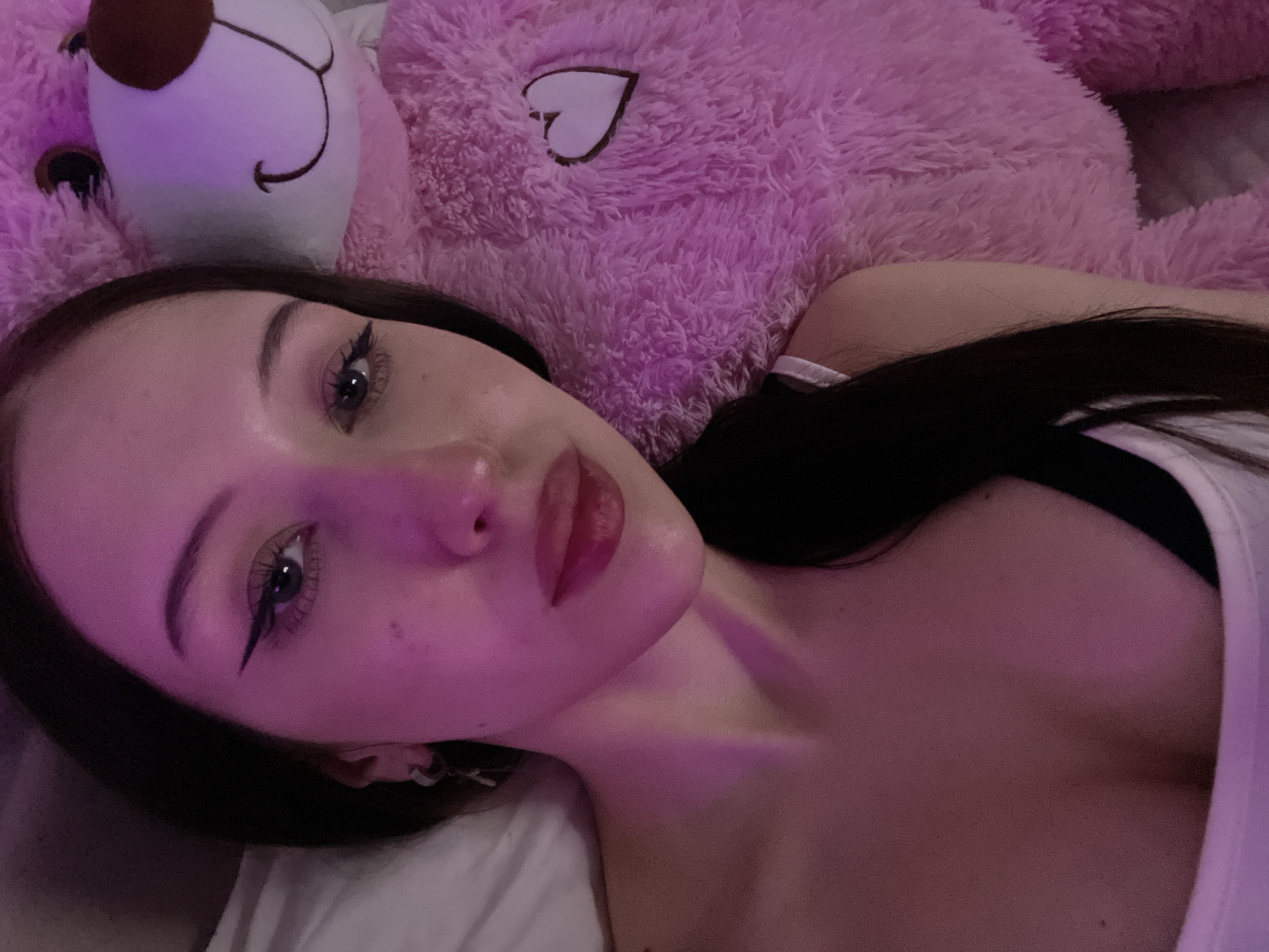 The scene is located in Votkinsk, Russia. A young adult female is depicted lying down, facing the camera directly. She has long dark hair, parted on her left, visible against a white pillow or bedding. Her eyes feature dark, winged eyeliner, and her lips are full with a reddish tint. A small hoop earring with a dangling element is visible on her left ear. She is wearing a white top, revealing her upper chest. To her right, a large, fluffy pink teddy bear is positioned, featuring a brown nose, a stitched smile, and a white heart appliqué with a dark border on its chest. The environment is illuminated by pink and purple ambient light, casting a glow on the subject's face and the teddy bear.