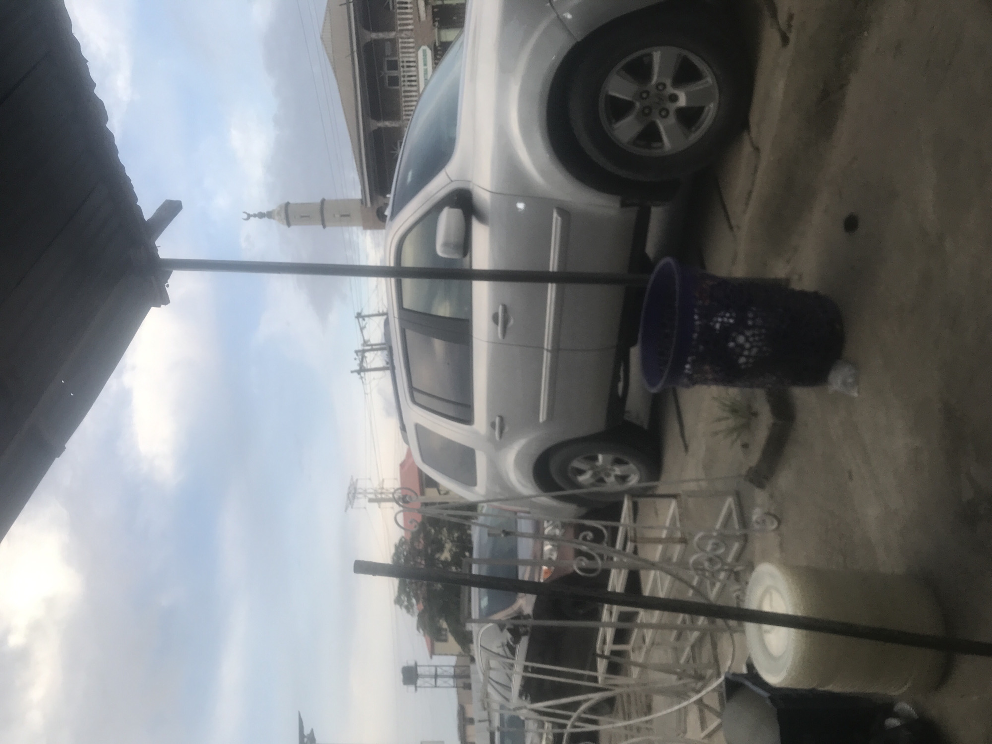 The image captures an outdoor scene, likely a street or parking area, under an overcast or cloudy daytime sky. In the midground, a silver sport utility vehicle (SUV), possibly a Honda Pilot, is parked on a paved surface. Adjacent to the SUV, on the ground, is a dark purple or blue trash can or bucket.
In the background, a prominent white minaret, topped with a distinct crescent moon symbol, stands tall, suggesting the presence of a mosque or Islamic architecture in the vicinity. Behind the SUV, part of a multi-story building with visible balconies can be seen. Overhead, utility poles and wires extend across the distant view.
The foreground features a white, ornate wrought-iron structure, possibly part of outdoor furniture or a decorative barrier, which partially frames the lower left and central areas of the image. To the right, a large, cylindrical roll of white material, resembling insulation or a fabric roll, rests on the ground. A dark-colored roof or awning is visible along the upper left edge of the frame. There are no people visible, nor is there any overt activity taking place. No discernible text is present in the image.