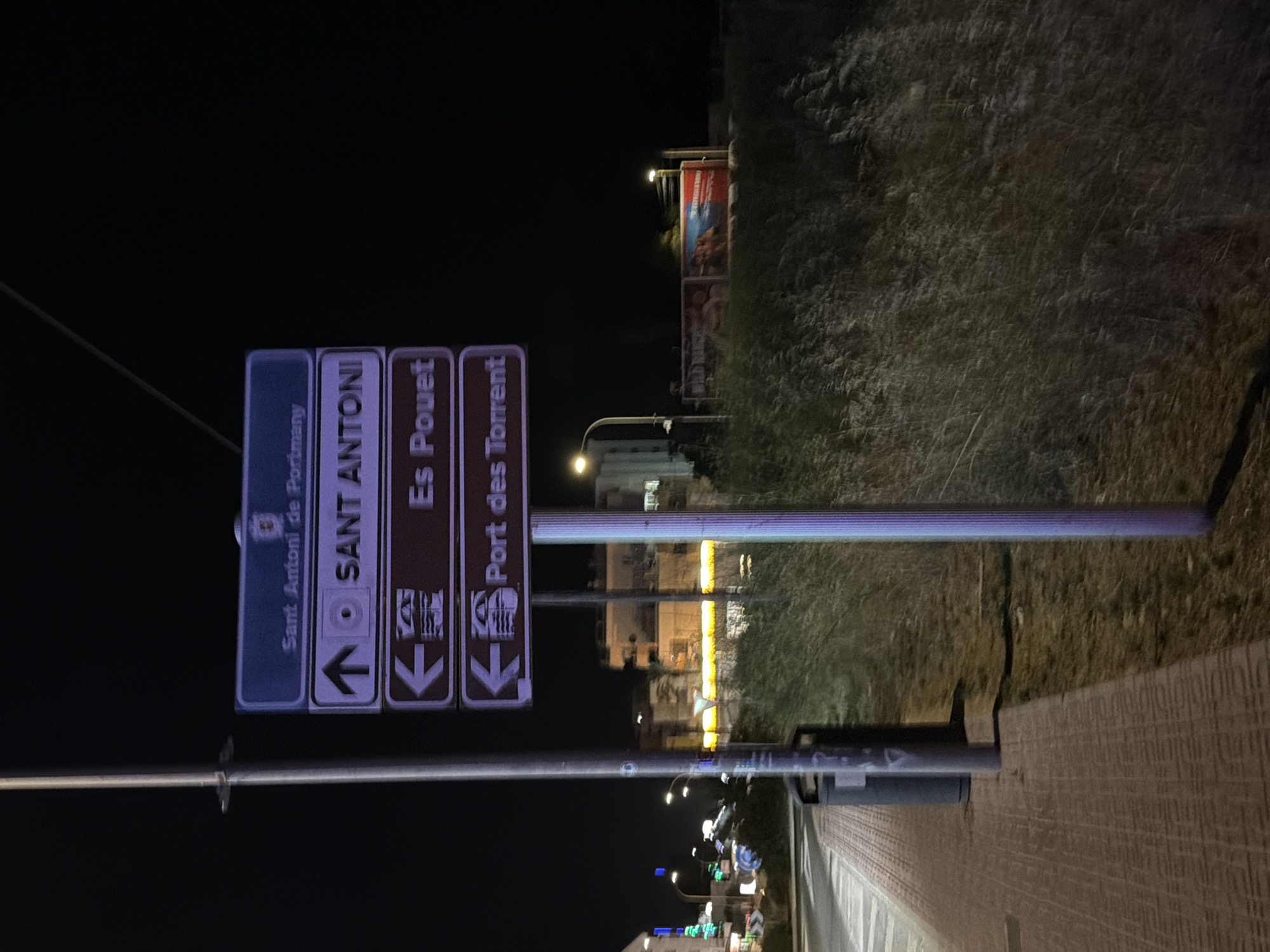 The image shows a street signpost in Sant Antoni De Portmany, Spain at night. The signpost has multiple signs indicating directions to different locations. The top sign, in a blue rectangle, reads "Sant Antoni de Portmany". Below this, a sign in a purple rectangle with a white arrow pointing upwards and a circular icon reads "SANT ANTONI". The next sign, in a brown rectangle with a white arrow pointing left and a circular icon, reads "Es Pouet". The bottom sign, also in a brown rectangle with a white arrow pointing left and a circular icon, reads "Port des Torrent". The signpost is illuminated by streetlights. In the background, there are buildings and vegetation, with some lights visible in the distance. A trash can is visible on the left side of the image, next to a sidewalk. The ground is paved with brick patterns.