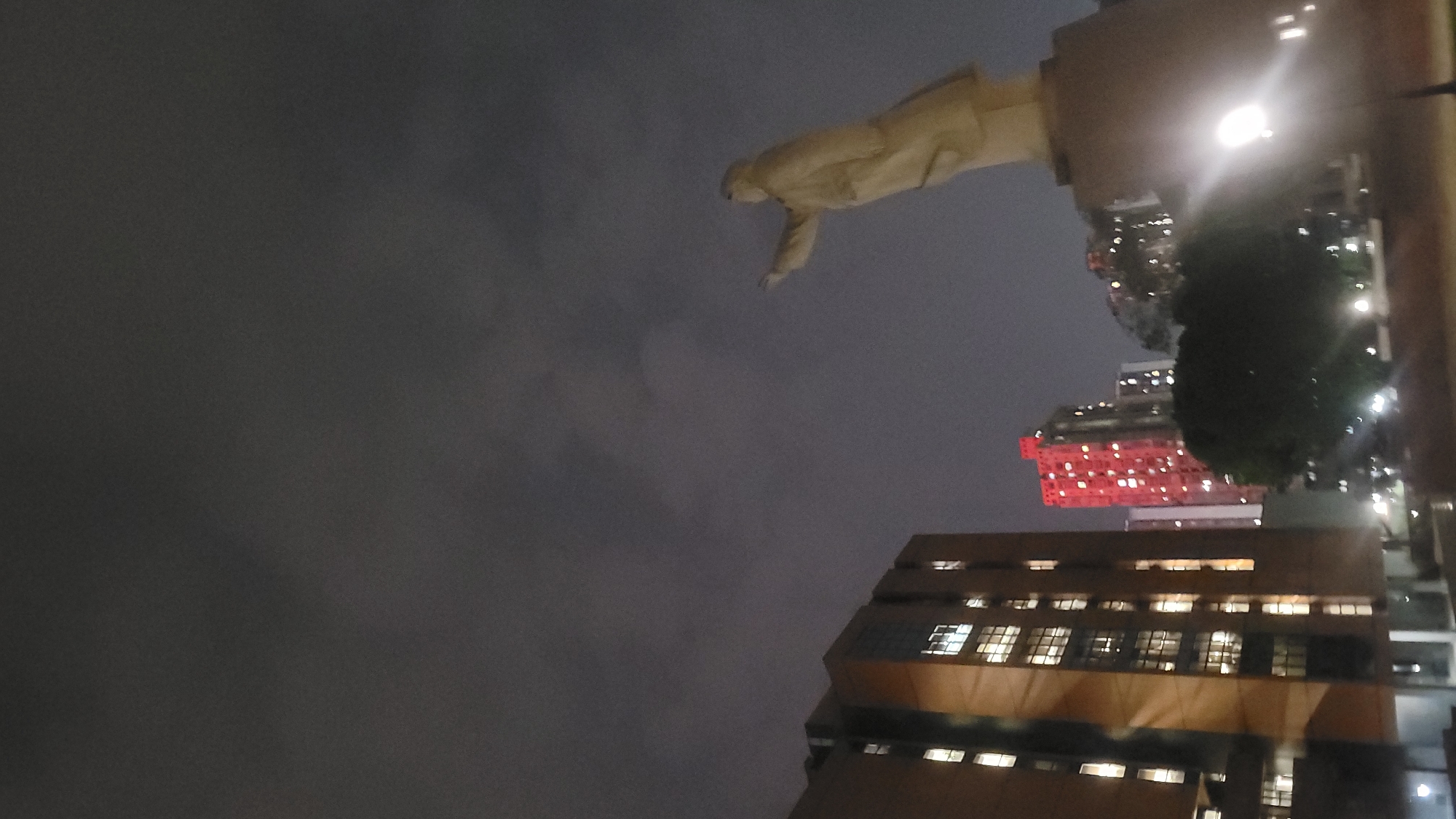The image, taken at night in Yuzhong District, China, is oriented at an angle. In the upper center, a light-colored statue stands atop a building. Below the statue, illuminated buildings are visible, including one with a distinctive red hue. The sky is dark and cloudy, filling a substantial portion of the frame. A large tree with dense foliage is positioned to the right of the red building.