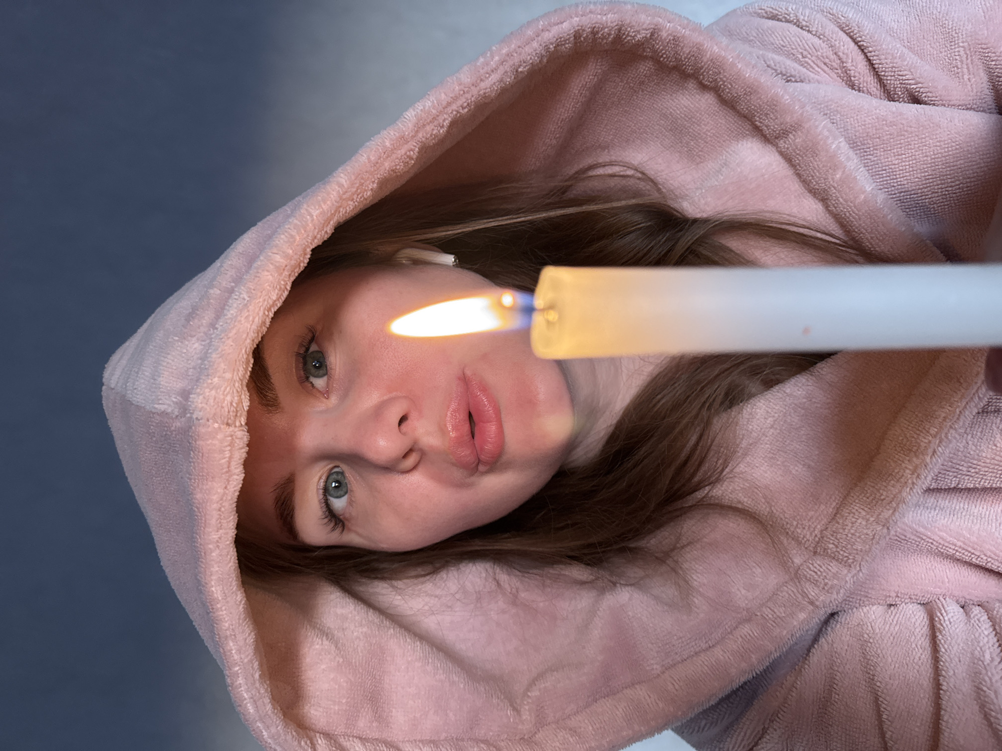 A woman in a pink hooded robe is holding a lit candle close to her face. She is looking at the camera with a slight pout. The background appears to be a wall with some light reflecting off it. The image does not provide enough information to determine if it was taken in Nestiukovo, Russia or any other specific location.