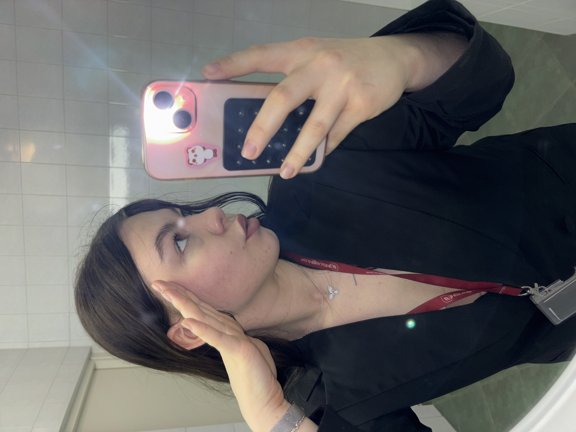 A young woman is taking a selfie in a bathroom mirror. She is wearing a black blazer and a silver necklace with a bird pendant. She has dark hair and is touching her face with her left hand. She is holding a pink phone with a bear sticker on the back. She is wearing a silver watch on her left wrist and a red lanyard with a white logo is visible around her neck. The bathroom has white tiled walls. The location is in the city of Perm, Russia.