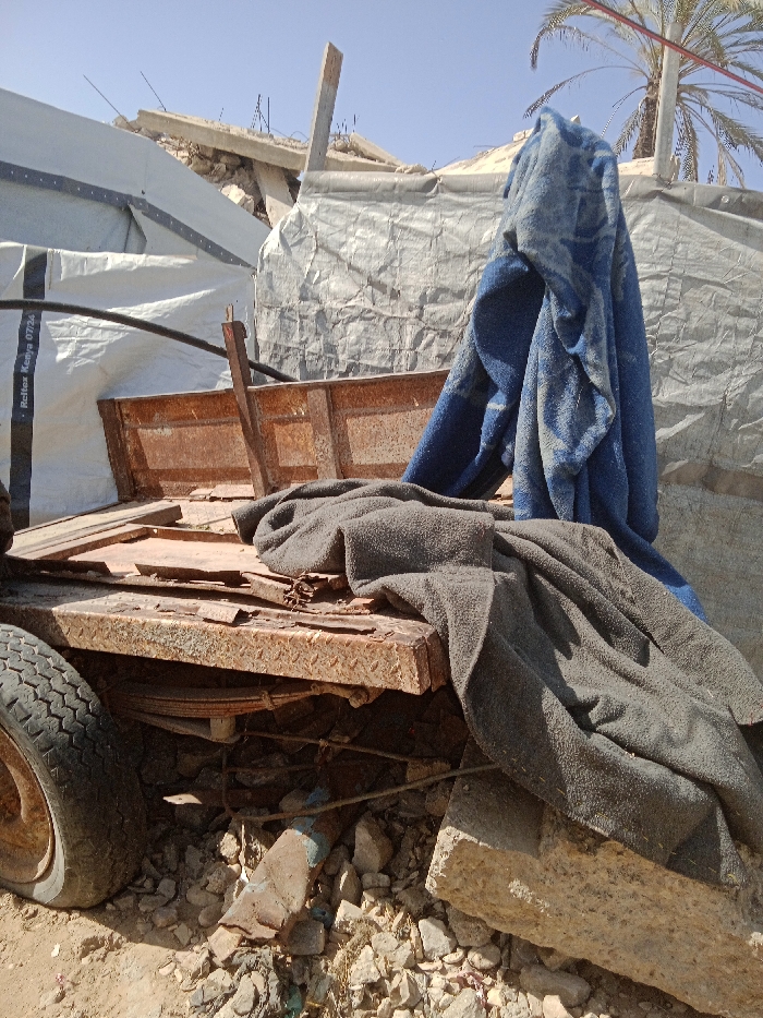 The image depicts a static outdoor scene in Khan Yunis, Palestinian Territory, characterized by makeshift structures and debris under clear daylight. In the foreground, the lower-left corner reveals a portion of a black rubber tire on a dirt ground scattered with small rocks and rubble. Central to the frame is a heavily rusted, orange-brown diamond plate metal trailer or wagon bed, partially covered by a dark grey fleece blanket. The trailer bed exhibits loose wooden planks and additional rusty metal components, including what appears to be a side panel. Beneath the trailer, leaf springs are visible amidst more rubble and a blue-painted metal fragment. A concrete block is positioned in the lower right, partially covered by the dark grey blanket.
Behind the trailer, a blue fleece blanket with a faint pattern is draped vertically. To the left of the blue blanket, a large, crinkled light-colored tarp or canvas is stretched across a structure, displaying text "Reflex Kenya 07/24". Above and behind this tarp, the remnants of a destroyed concrete structure are visible, featuring broken slabs, exposed rebar, and wooden supports against a clear blue sky. A palm tree with its trunk and fronds is visible in the upper right background. No individuals or active interactions are present in the scene.
