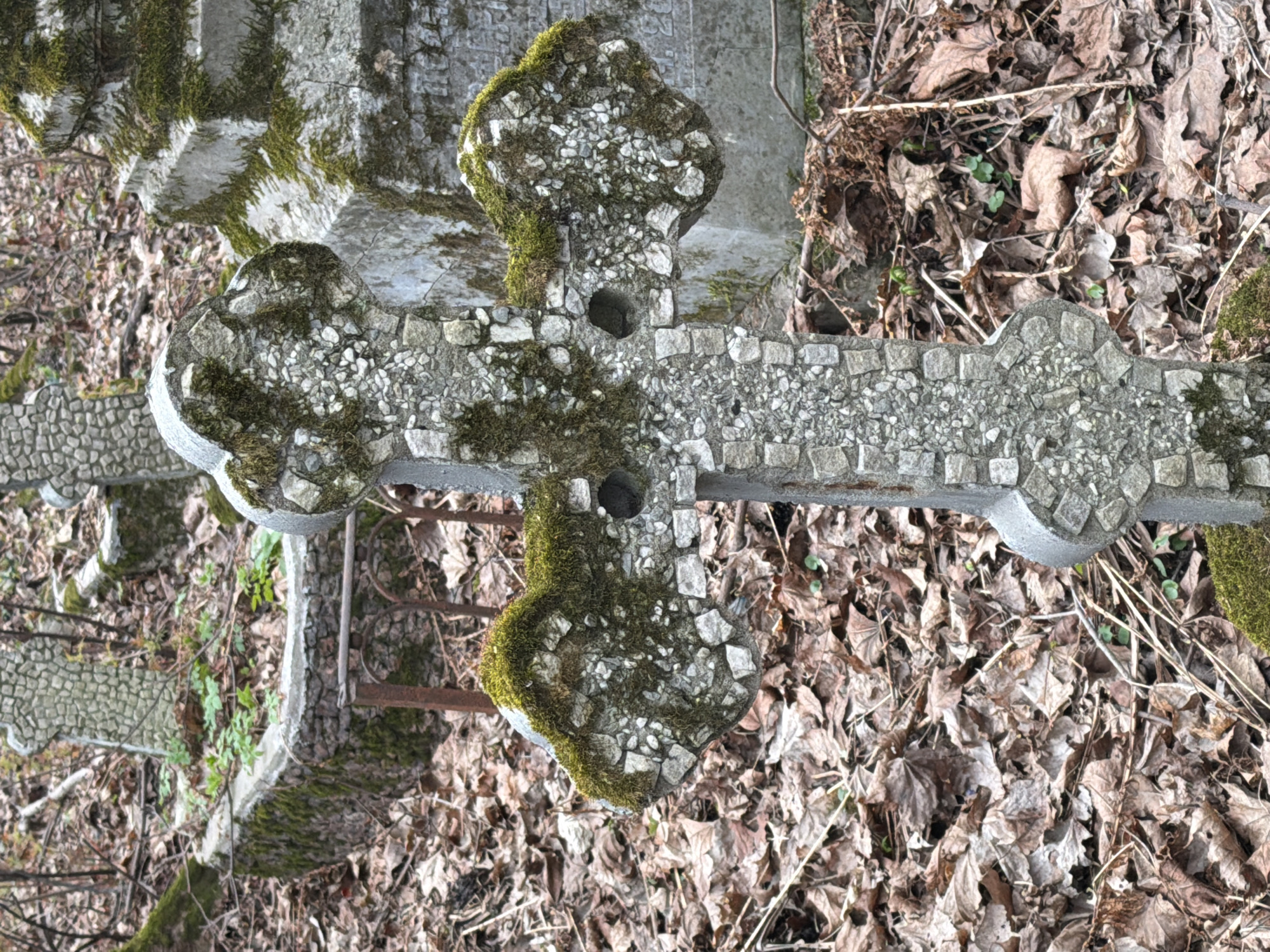 An overhead shot depicts a concrete cross, partially covered in moss, lying amidst fallen leaves. The cross is intricately designed with embedded stones. Its location is within Saint Petersburg, Russia.