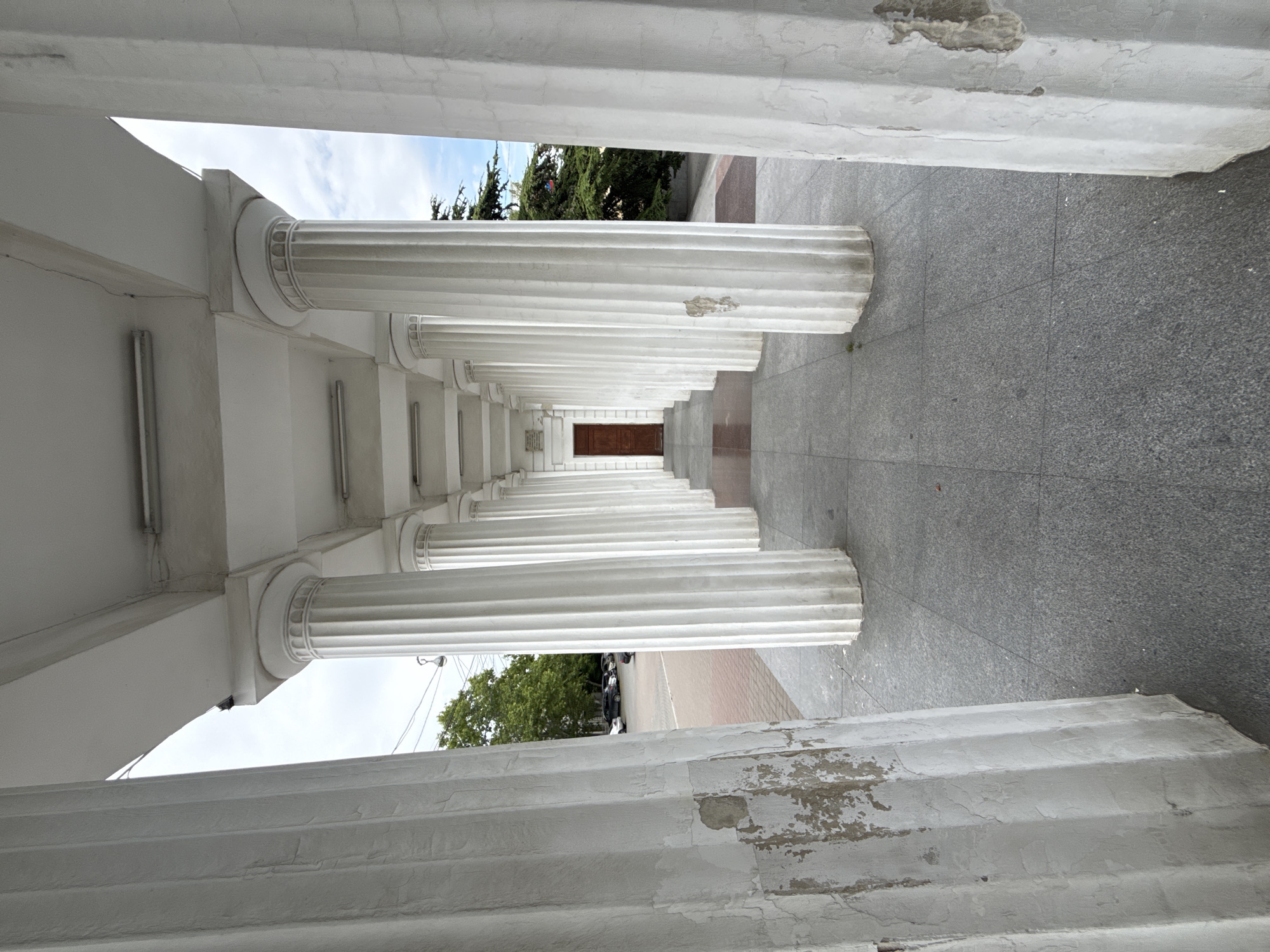 The image displays a perspective view along a white colonnade, looking from the exterior towards an interior closed door. The colonnade features two parallel rows of fluted, Doric-style columns, supporting a flat white ceiling. The two innermost columns in the immediate foreground on the left show significant peeling paint, exposing a dark, rough texture underneath. The remaining columns recede into the distance, appearing largely intact, with simple capitals. The ceiling houses several recessed rectangular fluorescent light fixtures. The walkway is paved with large, grey granite tiles. At the furthest point of the colonnade, a dark brown, closed double door is visible. To the left, beyond the columns, an urban street scene is evident, with parked cars, a large green deciduous tree, overhead power lines, and a streetlamp against an overcast sky. To the right, dark green coniferous trees are visible against a sky showing patches of blue and white clouds. No people are present in the scene. The location is in the city of Unknown, Unknown.