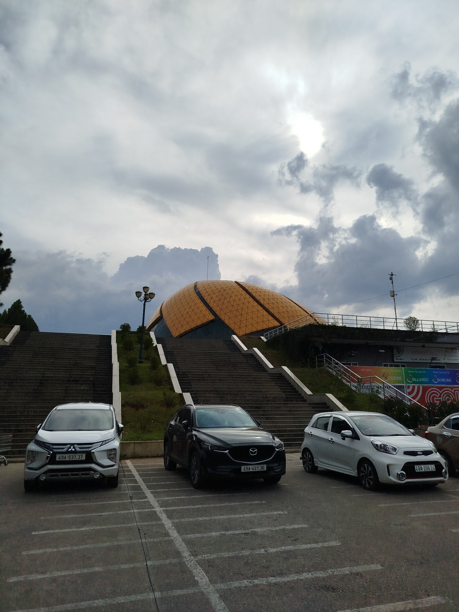 The image shows a parking lot in front of the Da Lat Lotus (Hoa Binh) Cultural Center in Da Lat, Vietnam. Three cars are parked in the foreground: a white Mitsubishi Xpander with the license plate "49A-537.37", a dark Mazda with the license plate "49A-427.36", and a white Kia Morning with the license plate "49A-208.24". Behind the cars, a wide set of concrete stairs leads up to the Lotus Center, an eye-catching golden dome structure. Along the right edge, a portion of the building behind the center is visible, showing signage for "KIDZONE", "BILLIARDS", and "BOWLING". The sky is overcast with dark, heavy clouds. A lamppost stands midway up the stairs. The architectural style of the Lotus Center suggests a post-modernist aesthetic.