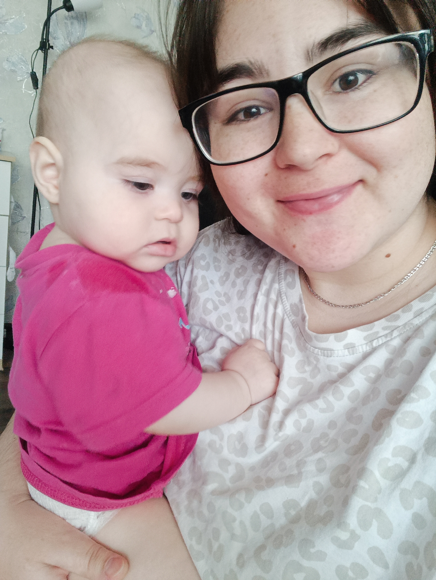 An indoor, close-up portrait shows a woman holding an infant in what appears to be a tender, casual moment. The woman, positioned on the right and smiling softly towards the camera, has dark hair and wears black-framed glasses. She is dressed in a light-colored t-shirt with a subtle grey and white animal print, and a thin silver chain necklace is visible around her neck.
The infant, held closely against the woman's chest on the left, has fair skin, light, sparse hair, and a neutral, slightly contemplative expression as it looks downwards and to the left. The baby is wearing a bright pink t-shirt and a white diaper.
The background is softly blurred, revealing a light-colored wall with a subtle patterned wallpaper, a black electrical cord or lamp part in the upper left, and a small portion of white furniture on the far left. The lighting is soft and even, suggesting daytime. No visible text is present in the image. This scene is likely set in a home in Noviy, Russia.