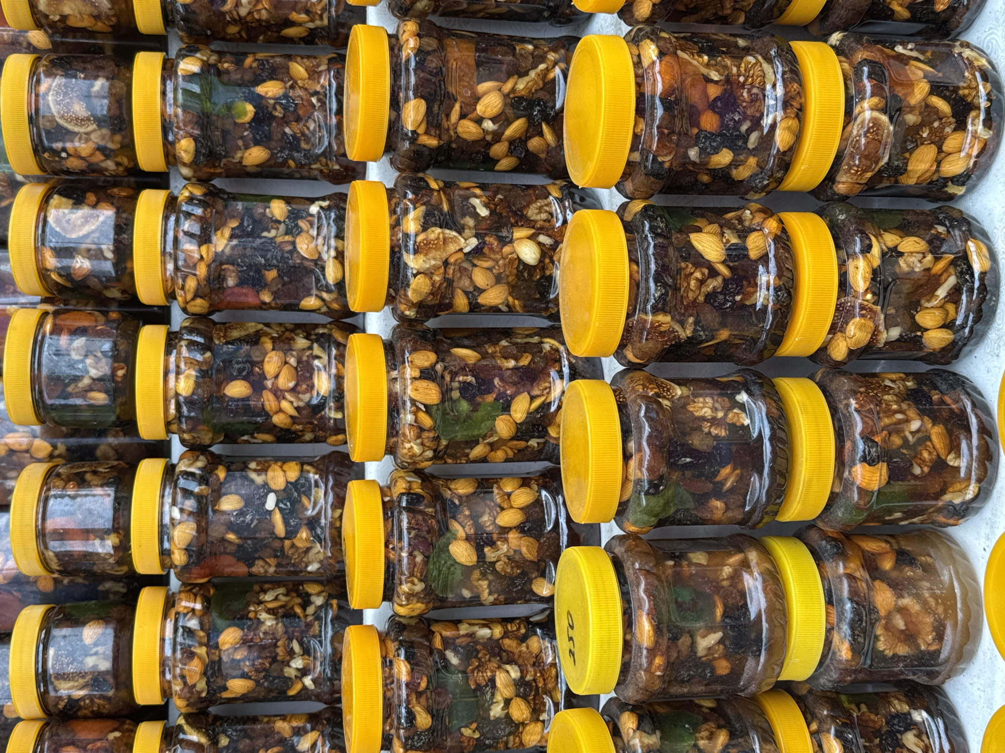 The image displays an overhead, close-up view of numerous transparent plastic jars, each filled with a dense mixture of nuts and dried fruits suspended in a dark, viscous liquid, likely honey or syrup. All jars are uniformly sealed with bright yellow screw-on lids and are arranged in neat rows and columns, creating a repetitive, grid-like pattern.
Inside the jars, distinct components are visible, including whole almonds, irregular pieces of walnuts, and slices of dried fruit, possibly figs or citrus, alongside other dark-colored dried fruits like raisins or dates. The contents appear rich and substantial.
The scene suggests a retail setting, such as a market stall or shop in Bishkek City, Kyrgyzstan, where these items are displayed for sale. The consistency in packaging and content points to a commercially prepared product. The lighting is even, allowing for clear visibility of the ingredients within the jars.
No human activity is depicted. On one of the bright yellow lids, towards the bottom right of the image, faint handwritten text "250" is discernible, which may indicate a price, weight, or product code. The overall impression is one of organized abundance, showcasing a local delicacy or health food product.