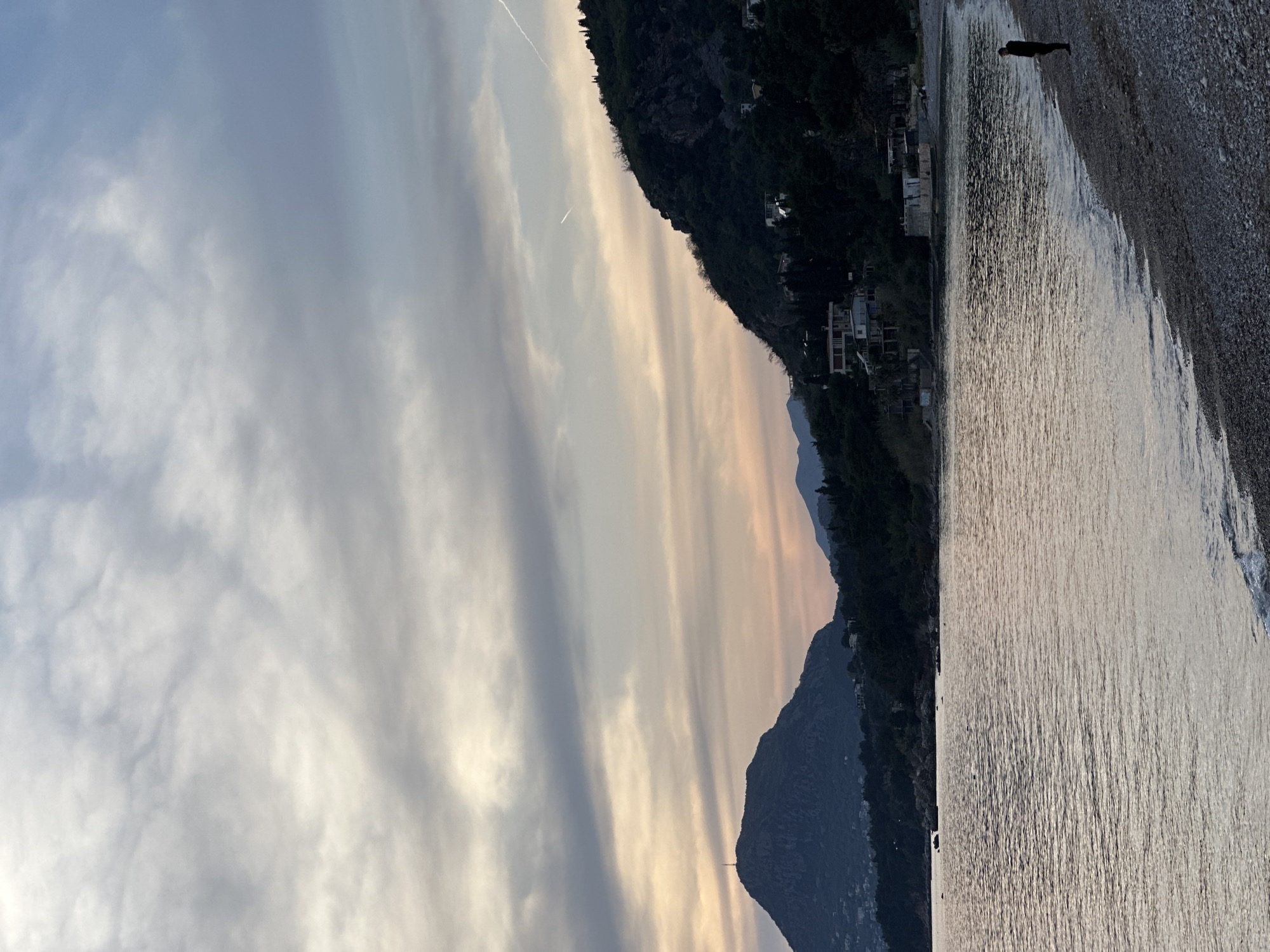 The image captures a coastline scene in Bar, Montenegro. The sky is overcast with layers of light grey and peach-toned clouds, and a contrail. A mountain range slopes down to the right, covered with dark green vegetation and buildings. A beach of grey pebbles is on the right, where the water meets the shore. A person is standing on the beach. The water reflects the light, appearing silver and textured.