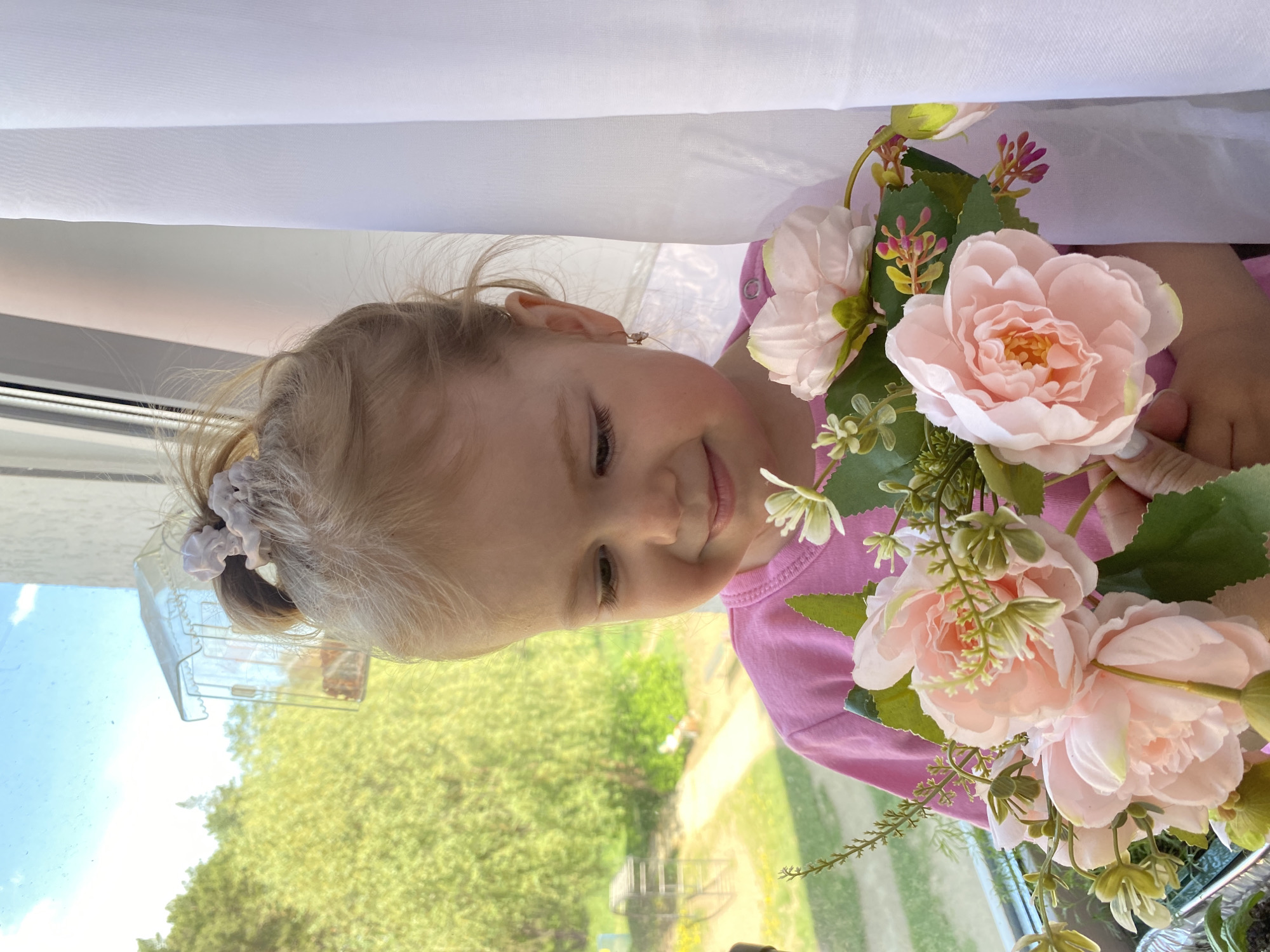 A young girl with fair hair, partially gathered with a pink hair tie, is holding a bouquet of artificial pink flowers. She is wearing a pink long-sleeved shirt. She is smiling slightly and looking down towards the flowers. She has an earring in her left ear. She is positioned in front of a window, through which trees and the sky are visible. A white sheer fabric is draped above her, partially covering the top of the frame. Behind the girl, a glimpse of the city of Chaikovsky, Russia is seen through the window.