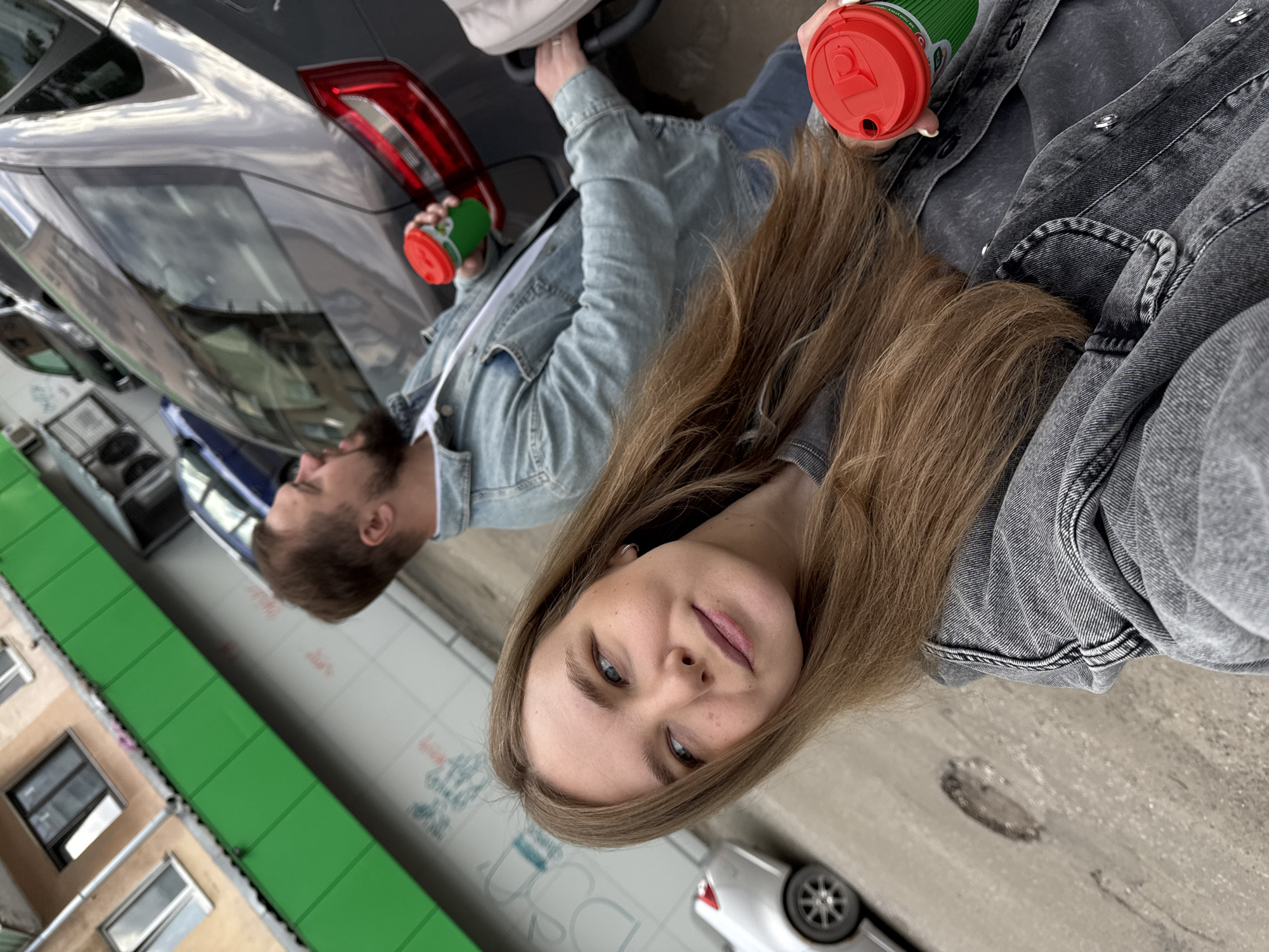 The image depicts two individuals standing near a gray car on a street in Vladimir, Russia. A woman with long brown hair, wearing a gray denim jacket, is in the foreground, facing the camera and holding a green coffee cup with a red lid. Behind her, a man with short brown hair, wearing a light blue denim jacket, is visible, also holding a similar coffee cup. In the background, there is a building with windows and a green structure, and a white car is parked on the street. The scene appears to be a casual, everyday moment captured on a city street.