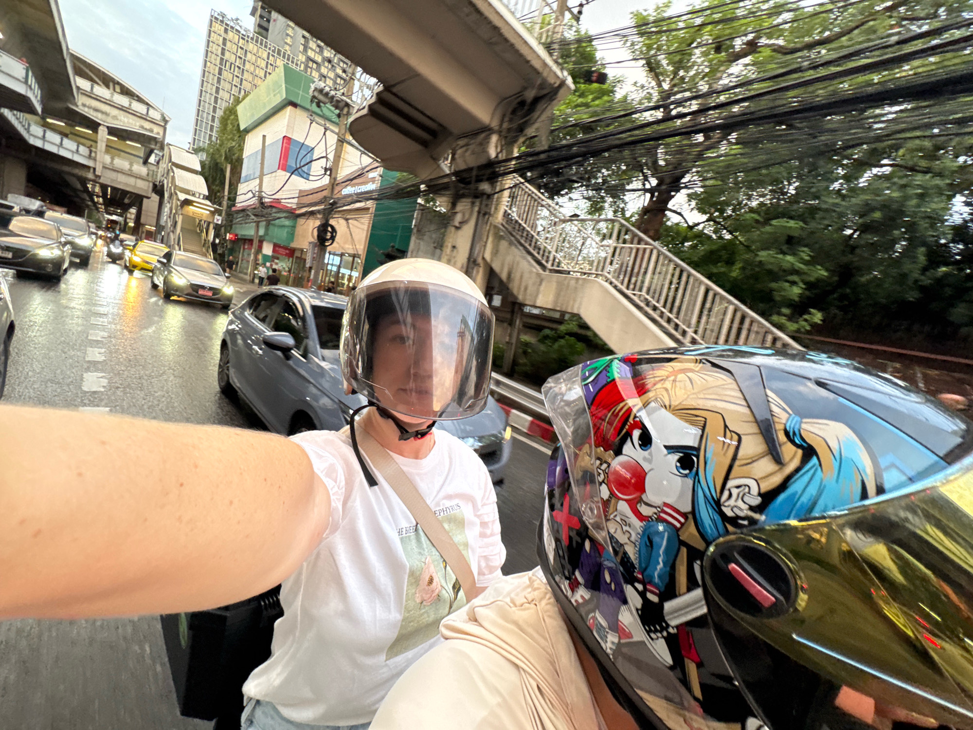 Two individuals are riding on a motorcycle in an urban environment in Bangkok, Thailand. The person in the foreground is wearing a helmet with a cartoonish design featuring a character with blue hair and a red nose. Their arm is visible, resting on the motorcycle. The person behind them is wearing a white t-shirt with a floral print and a beige strap across their chest, and a clear helmet. The motorcycle is on a wet road, suggesting recent rain, and is surrounded by other vehicles including cars and a yellow taxi. In the background, there is elevated infrastructure, likely part of Bangkok's public transportation system, such as an overpass or a BTS Skytrain line, along with buildings and trees. The overall scene depicts a typical street view in Bangkok during daylight.