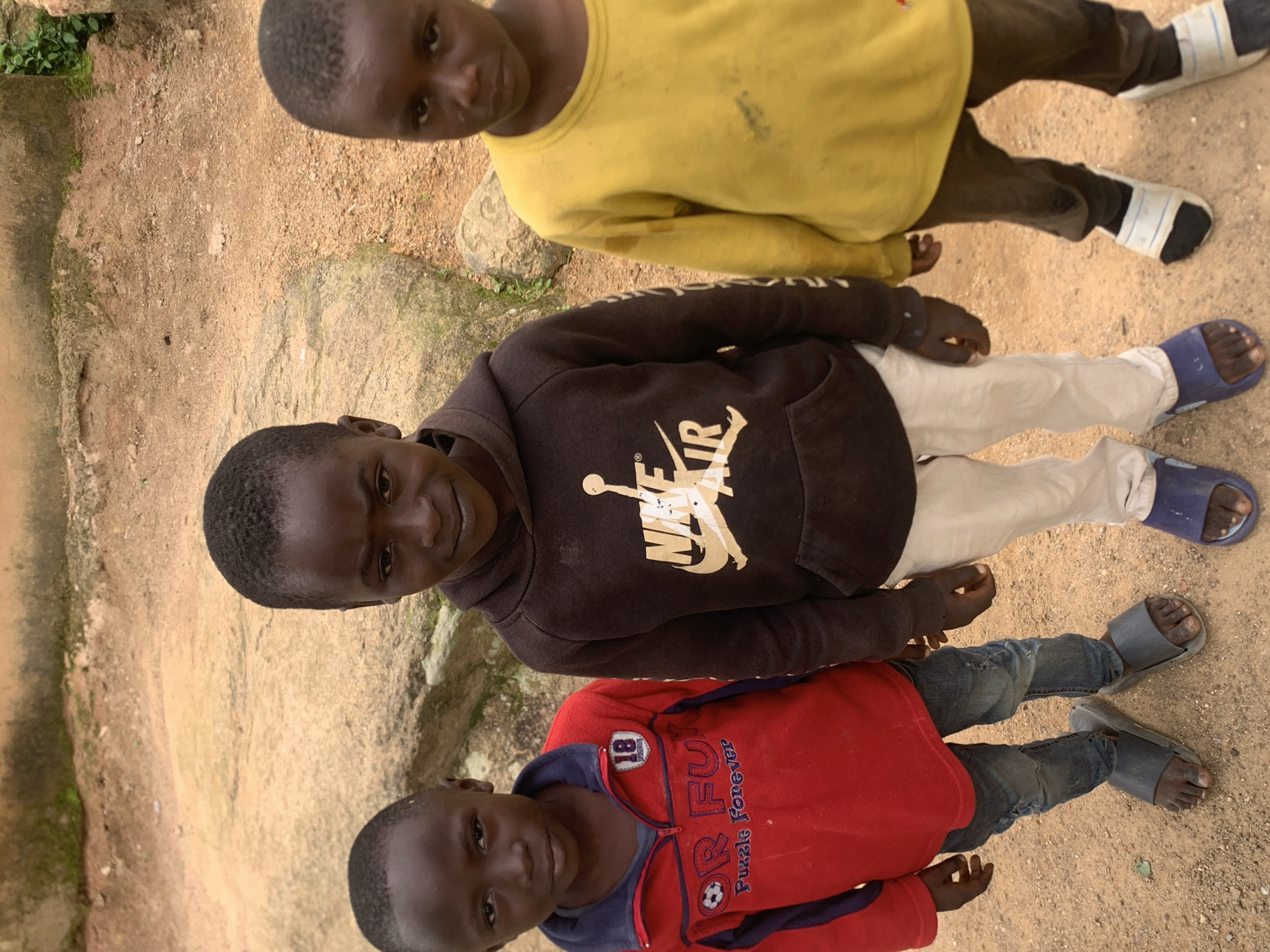Three young boys stand outdoors in Jos, Nigeria. The boy on the left wears a red jacket with blue trim and the text "RFU PUZZLE FOREVER" with a soccer ball emblem. He is also wearing blue jeans and dark grey slip-on sandals. The boy in the center is wearing a dark brown hoodie with a large white Nike Air Jordan logo on the chest and light beige pants. He is wearing blue slip-on sandals. The boy on the right is wearing a yellow long-sleeved shirt and dark pants, along with white and black slip-on sandals. They are standing on a dirt and gravel ground with rocks and sparse vegetation visible in the background. The overall scene suggests a simple, possibly rural or less developed urban environment.