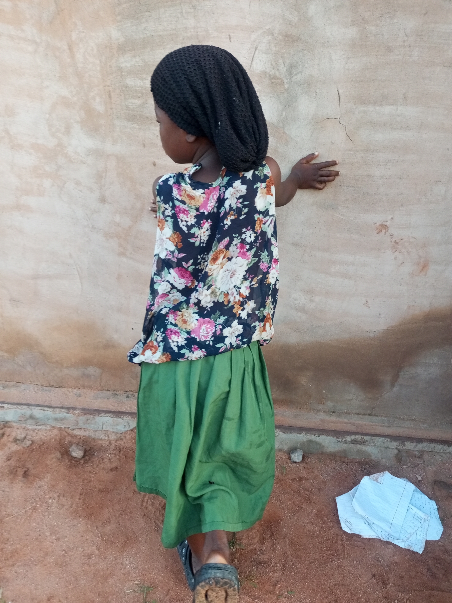 In Tempure, Nigeria, a young person wearing a black head covering, a floral patterned top, and a green skirt stands with their back to the camera, leaning against a textured, light brown wall. Their right hand is extended to touch the wall, with fingers spread. The ground is a reddish-brown earth, and a stack of white papers with writing is visible on the ground to the right of the person. The lighting suggests it is daytime, and the overall environment appears to be outdoors, possibly in a rural or less developed urban area.