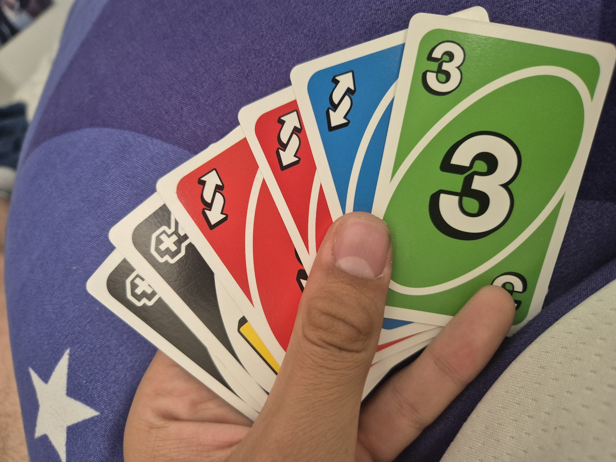 A hand holds six UNO cards, with a green "3" card facing upwards. From left to right, the cards appear to be black "Draw Two", black "Draw Two", red "Reverse", red "Reverse", blue "Reverse", and green "3". The cards are held over a blue and white patterned surface.