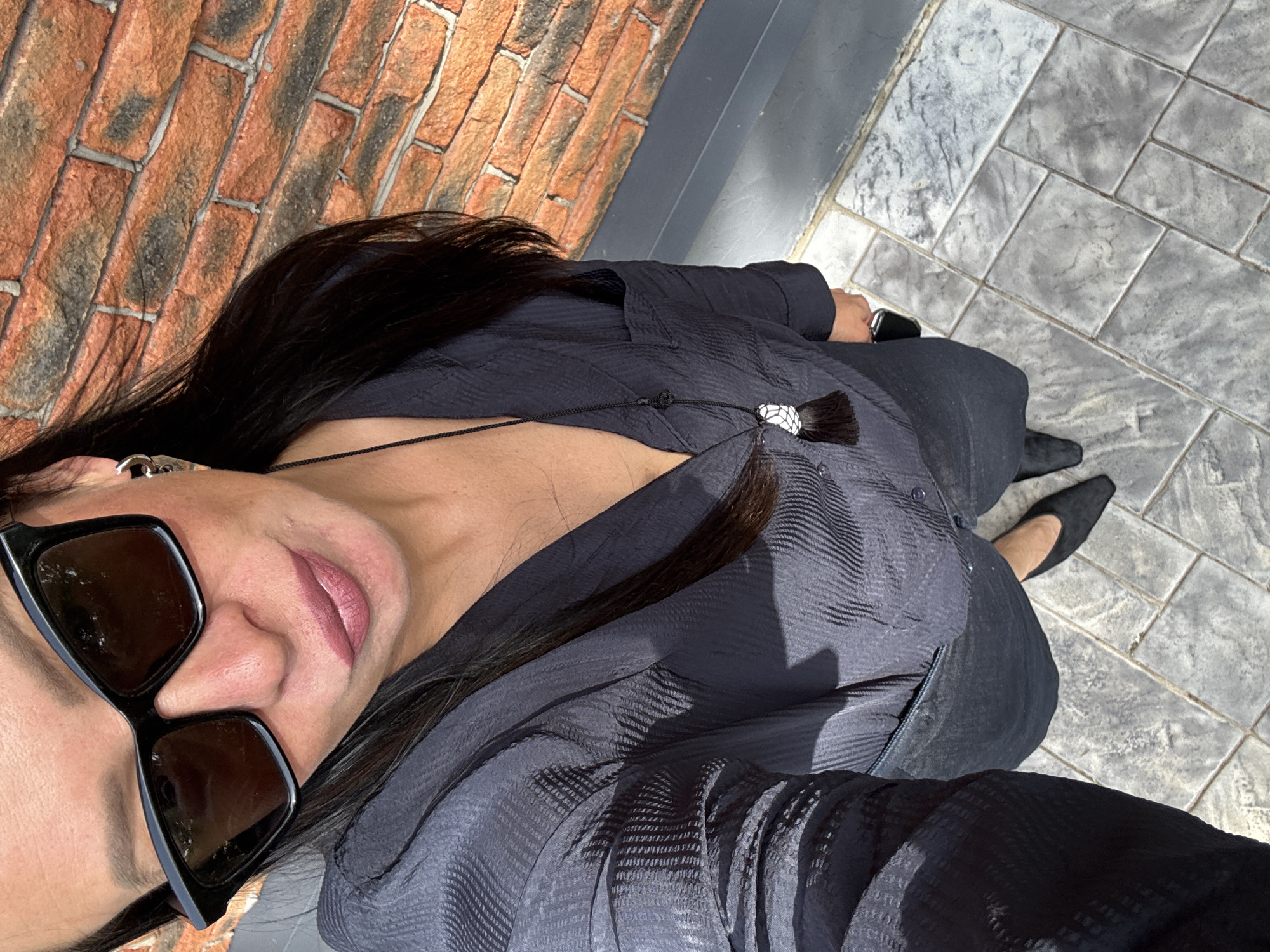 A woman with dark hair is posing for a selfie outdoors. She is wearing sunglasses and a dark-colored outfit. She is standing on a stone-paved surface, with a brick wall visible in the background. The location could be Chaikovsky, Russia.