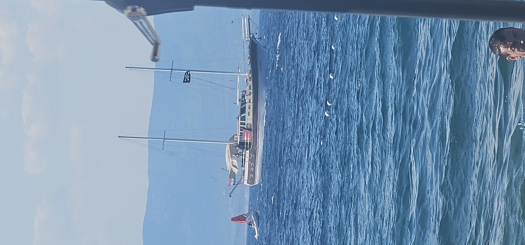 A wide shot captures a vast expanse of blue water under a bright, clear sky. In the mid-ground, a large, traditional wooden sailing boat with two masts dominates the scene, its sails partially furled. A Croatian flag flies from the bow of the boat. A black flag with a skull and crossbones is visible on the main mast. Several people are standing on the deck of the sailboat. To the left of the sailboat, a smaller dinghy with a person aboard and a red sail is also on the water. In the foreground, a line of white buoys floats on the surface of the water, connected by a rope. The far background features hazy blue mountains. In the lower right corner of the frame, the head and shoulders of a man are visible, partially submerged in the water. The scene appears to be set in a coastal area.