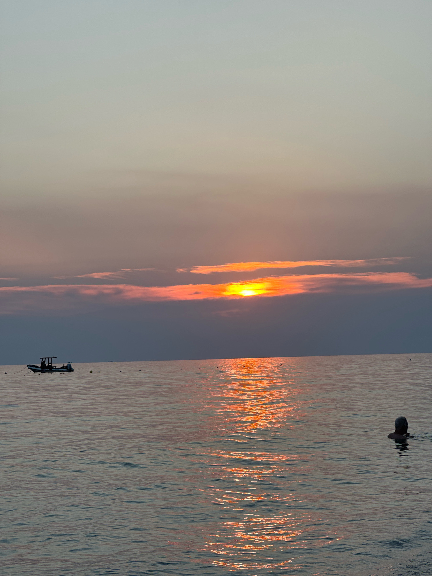This is a landscape photograph of a body of water at sunset. The sky is a gradient of pale blue to muted orange and pink, with a band of darker clouds across the middle where the sun is setting. The sun itself is a bright orange disc peeking through a gap in the clouds. The water is calm, with gentle ripples that reflect the colors of the sunset. In the distance, on the left side of the frame, a small boat with a dark hull and white superstructure is anchored. There are also several small buoys scattered in the water. On the right side of the frame, a person is swimming in the water, with their head and shoulders visible. The overall mood of the image is peaceful and serene.