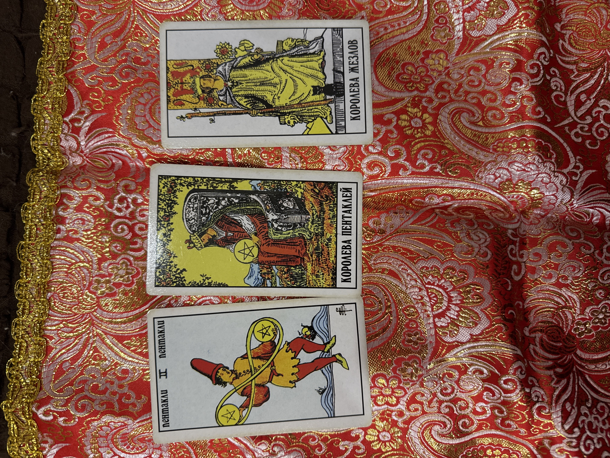 The image shows three tarot cards laid out on a red and gold patterned fabric. The top card is "Queen of Wands" ("Королева жезлов"), the middle card is "Queen of Pentacles" ("Королева пентаклей"), and the bottom card is "Two of Pentacles" ("пентакли II"). The location is not discernible and the image may not be in Sochi, Russia. There are no people visible.