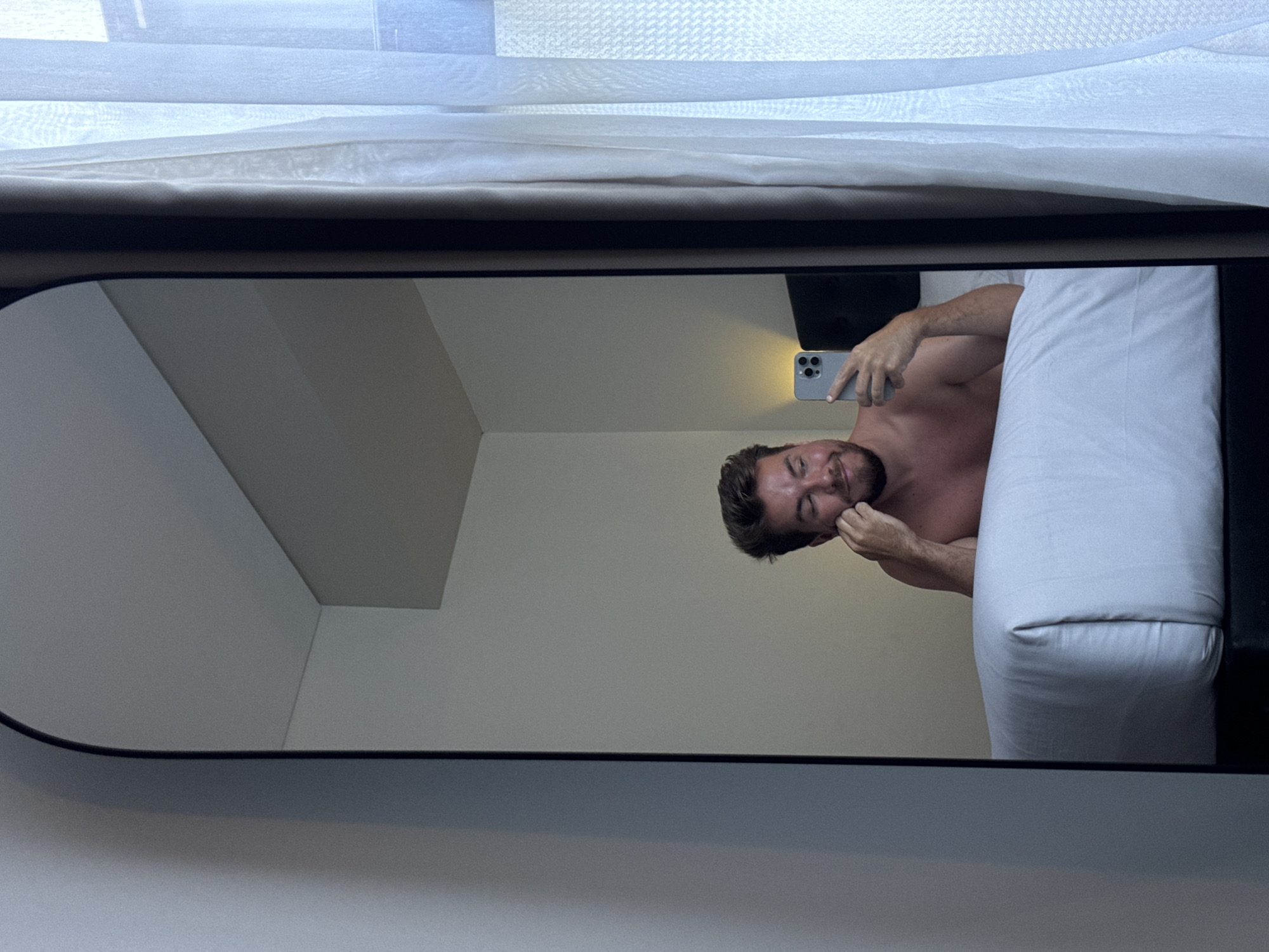 A shirtless man in a room is reflected in a mirror. The man holds a cell phone in his right hand and touches his face with his left. To the right of the man is a white pillow. The room's walls are pale. The top of the mirror reflects a white curtain. The location is North Kuta, Indonesia.