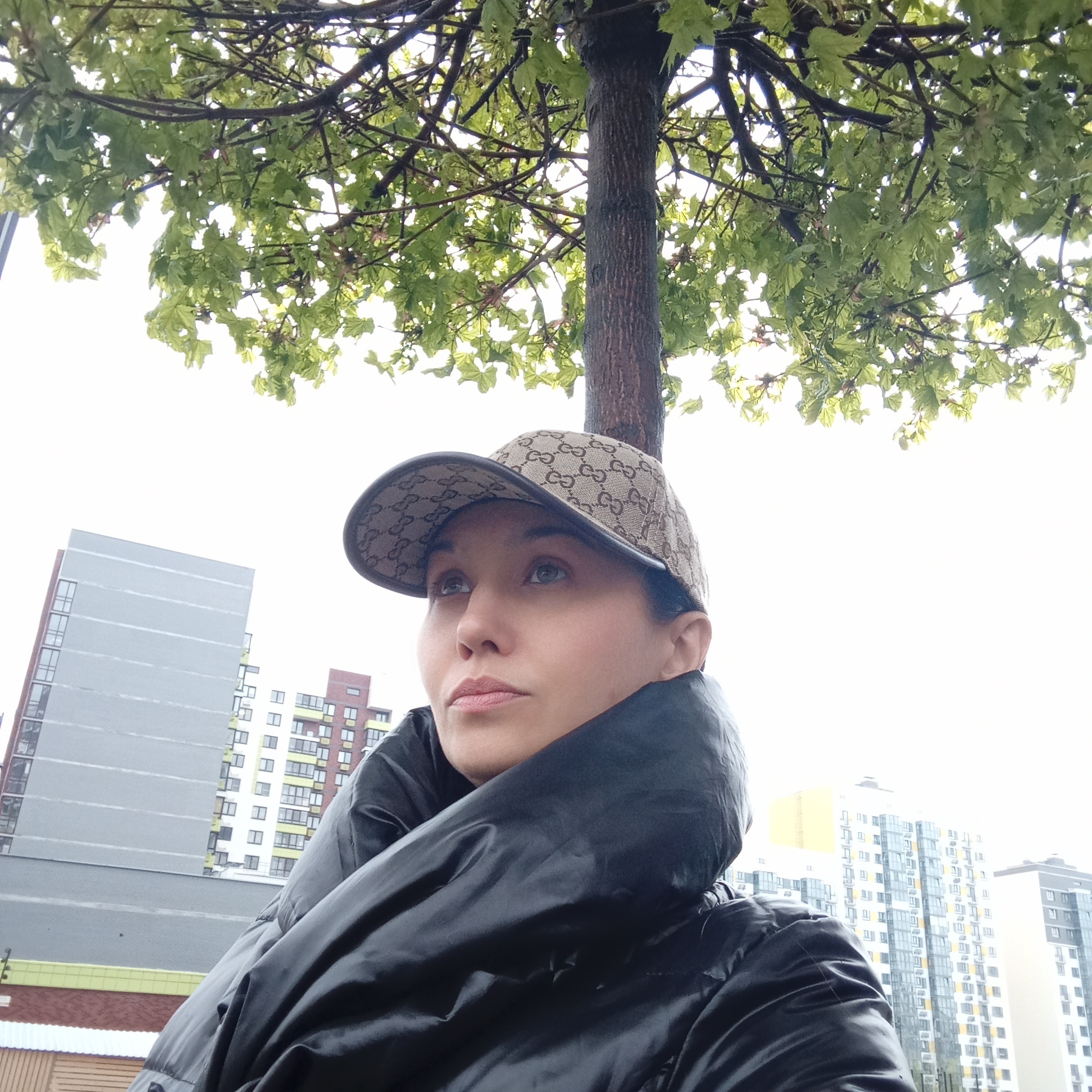 The image shows a woman in Izhevsk, Russia. She is wearing a Gucci baseball cap and a large, black, puffy coat. She is looking up and to the left of the frame. A tree trunk is directly behind her head, with green leaves visible in the upper portion of the frame. In the background are several tall buildings characteristic of the city. The sky is overcast.