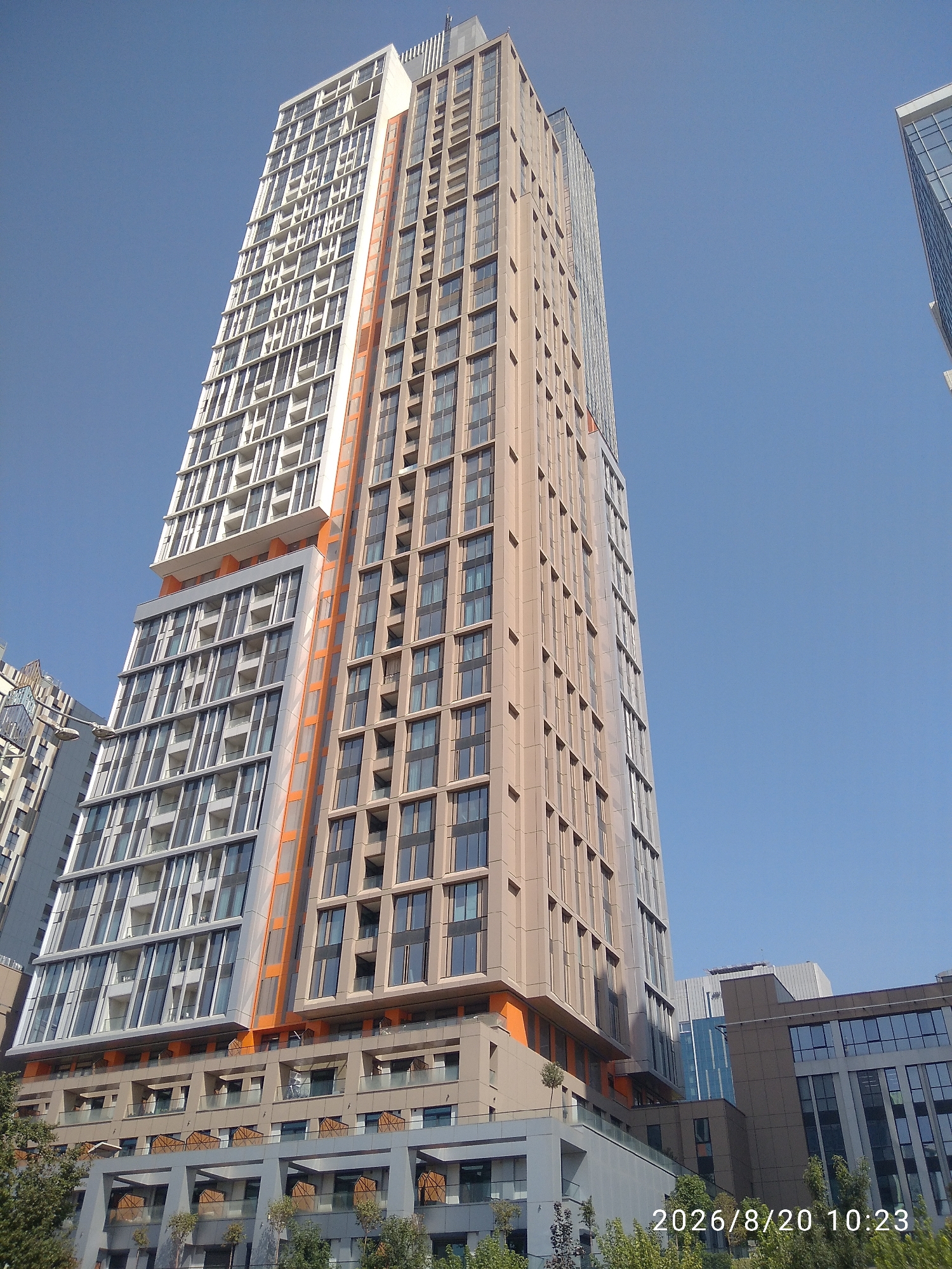 A low-angle shot captures the modern architecture of a skyscraper in Tashkent, Uzbekistan, under a clear blue sky. The skyscraper features a distinctive design with a light-colored, patterned facade on the upper floors and a beige facade on the lower sections, accented with orange detailing. To the right, another modern building with a glass facade is partially visible. In the foreground, trees and the lower levels of buildings with balconies and windows are visible. A date and time stamp, "2026/8/20 10:23," is present in the lower right corner of the image.